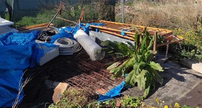 Pila de artículos desechados cubiertos con una lona azul; incluye metal, madera y una planta, al aire libre.