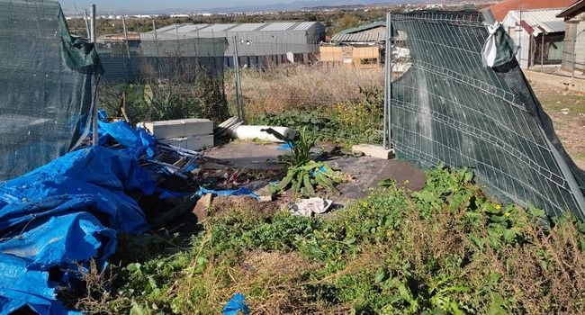 Un área exterior desordenada con una lona azul, maleza crecida y una cerca de alambre.