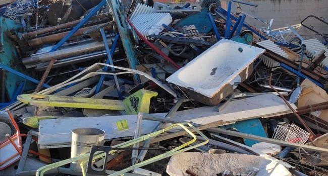 Montón de basura metálica que incluye una bañera, piezas de bicicleta y muebles, al aire libre.