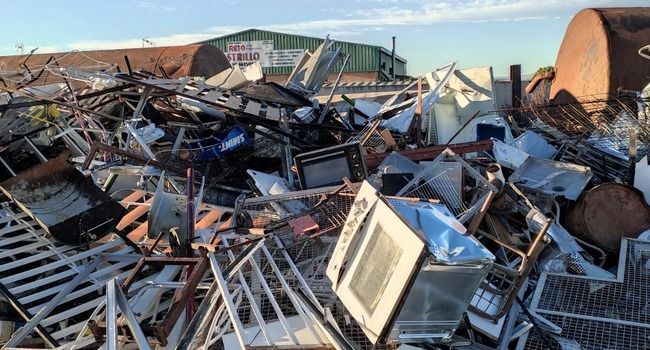 Pila de chatarra en una instalación de reciclaje; incluye electrodomésticos, marcos y un tanque grande.