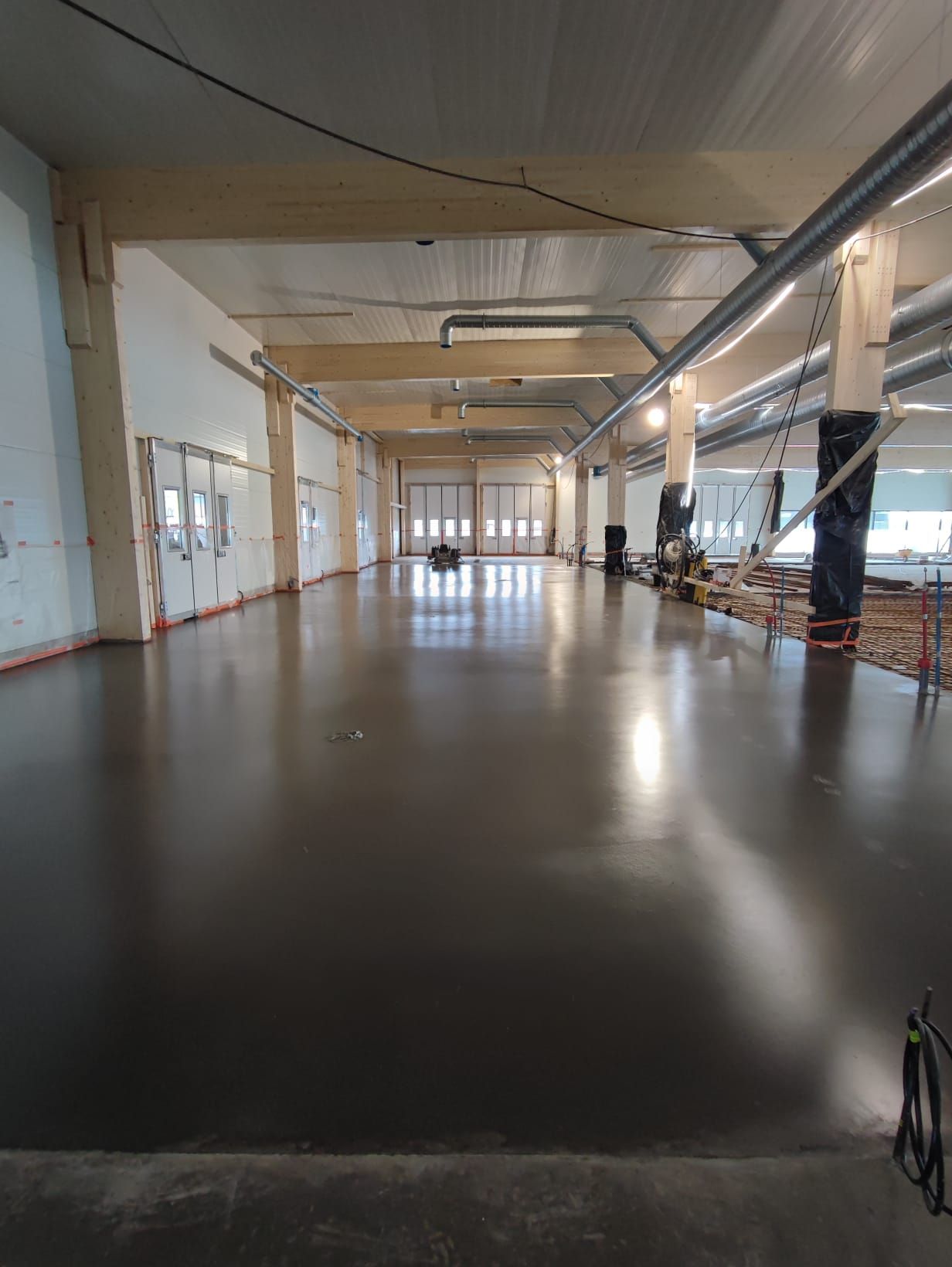 Leveä, tyhjä teollisuustila, jossa on tumma, heijastava lattia ja kattopalkit.