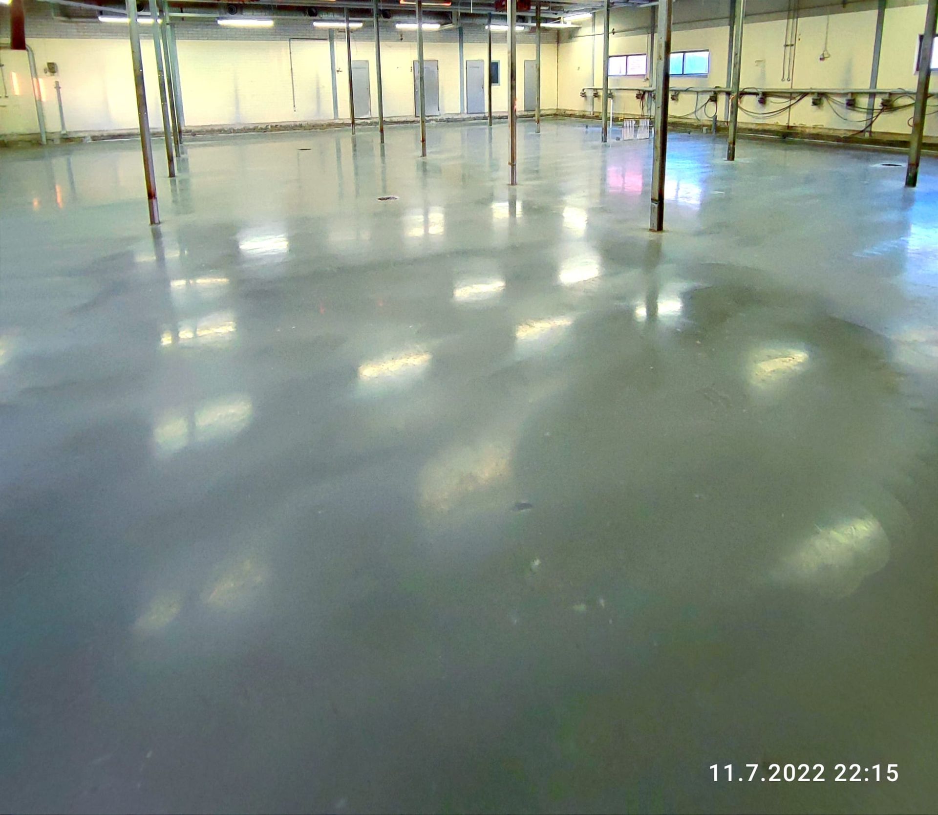 Tyhjä teollisuustila, jossa on kiiltävän harmaa lattia, metalliset tukipalkit ja kattovalot.