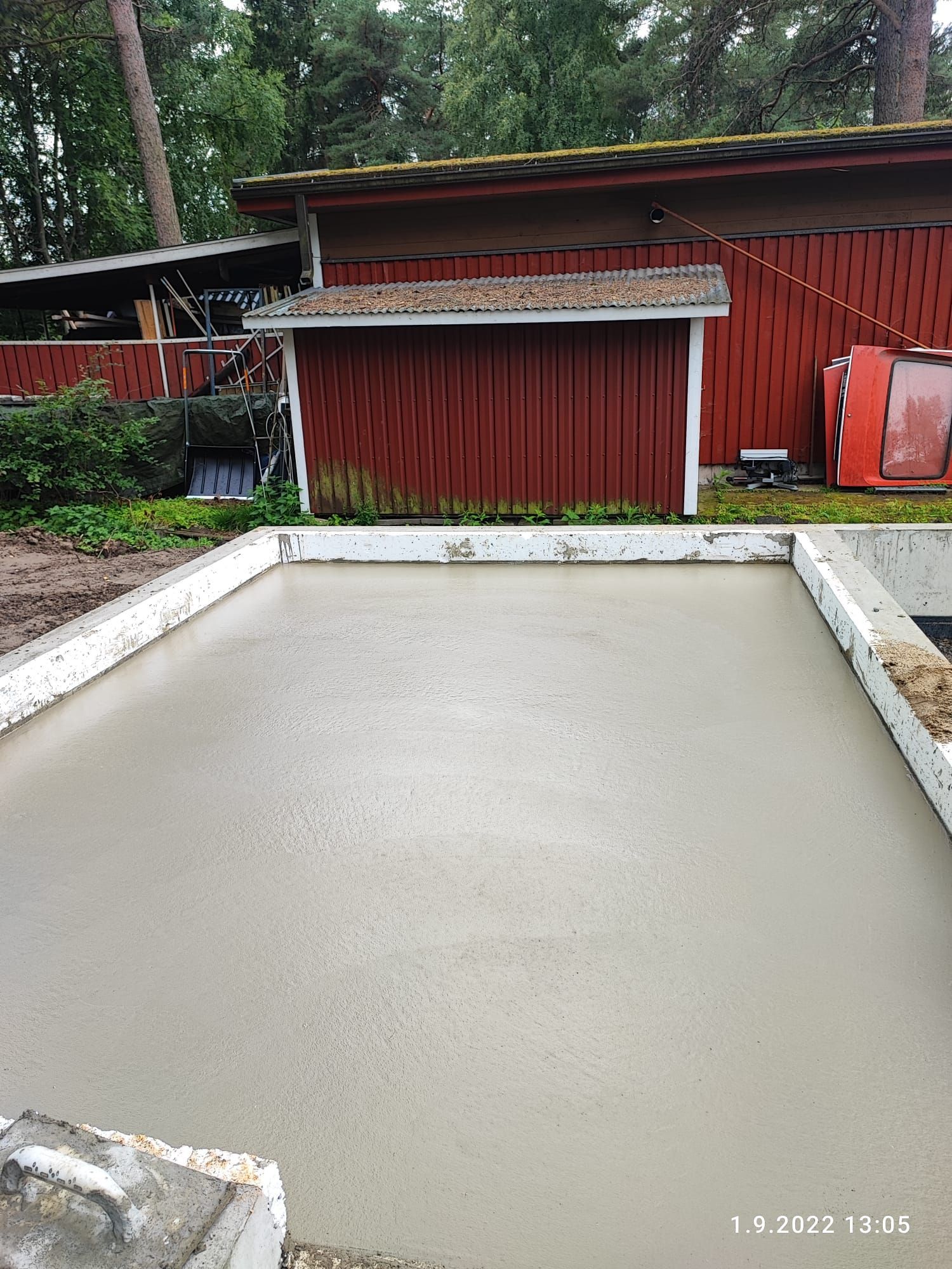 Vastavalettu betonipinta puukehyksen sisällä punaisen rakennuksen ja puiden edessä.