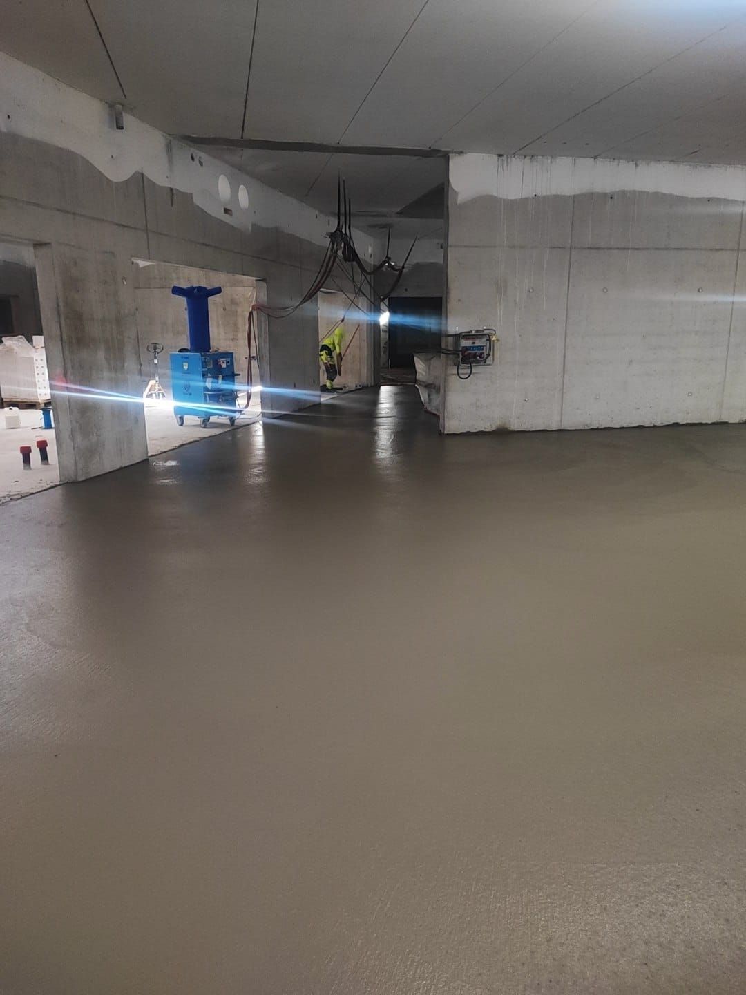Vastavalettu betonilattia keskeneräisessä rakennuksessa. Rakennustyöntekijä taustalla, koneet näkyvissä.