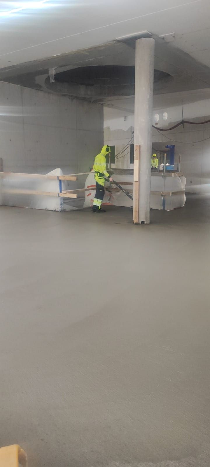 Heijastinvaatteissa oleva henkilö tasoittaa märkää betonilattiaa keskeneräisessä rakennuksessa.