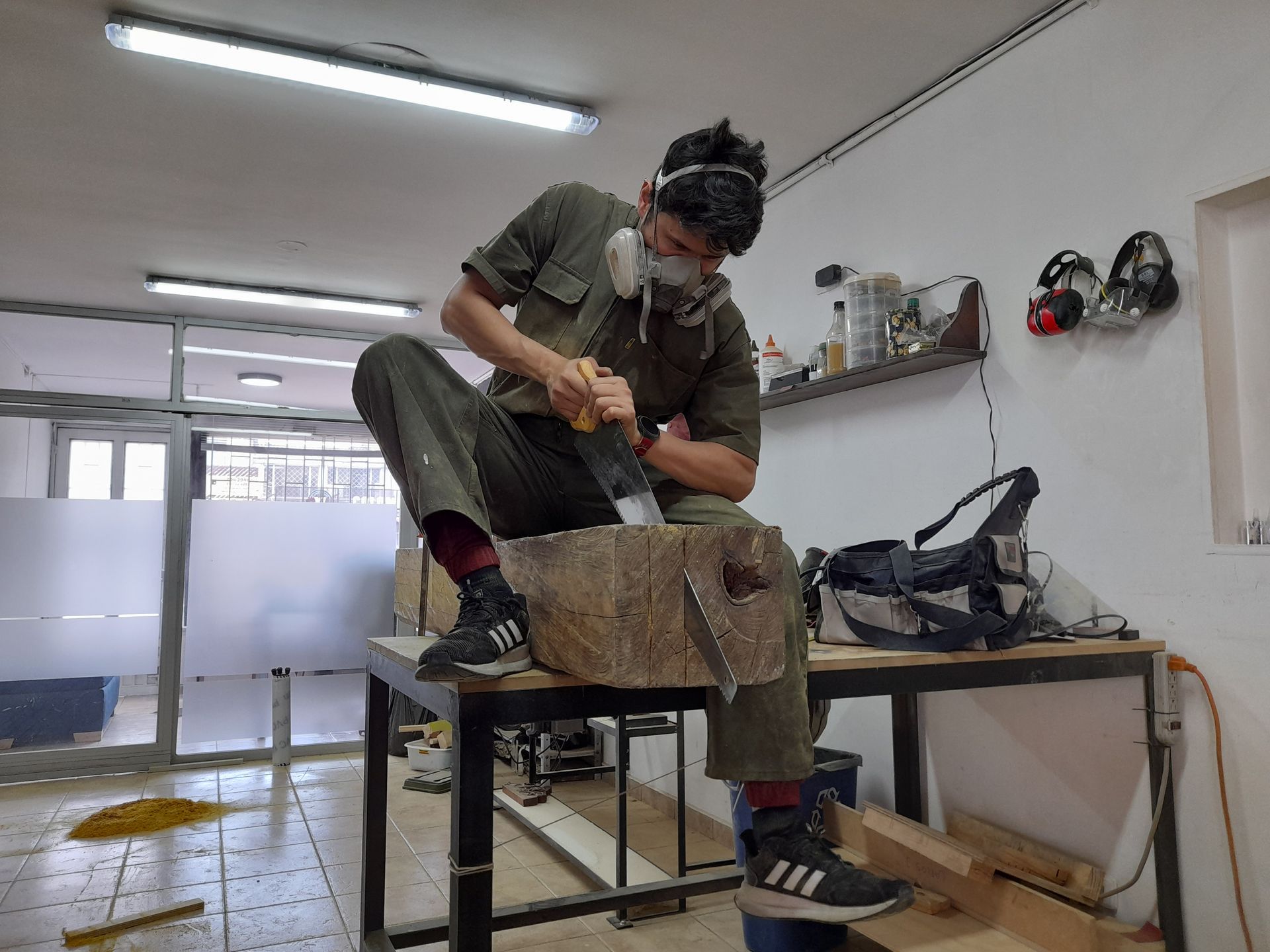 Persona tallando madera, con mascarilla, en un taller. Sentada en una mesa de trabajo, utilizando un cincel.