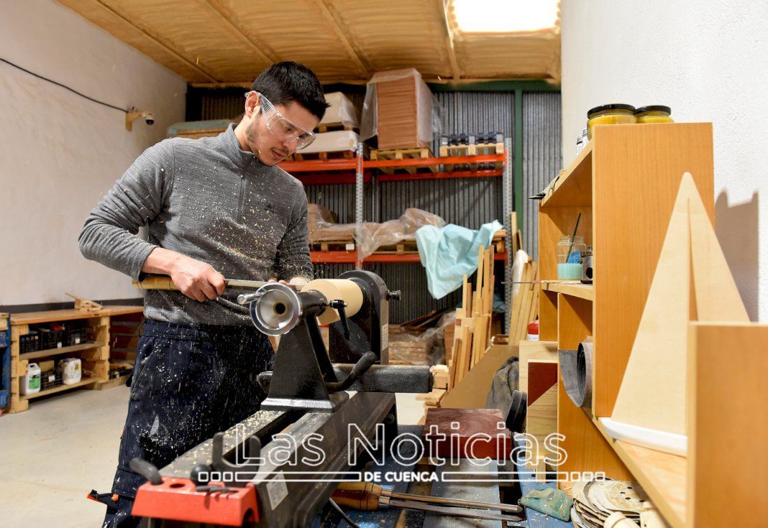 Hombre en un taller utilizando un torno, con gafas de seguridad y delantal. Al fondo se ven estanterías y herramientas