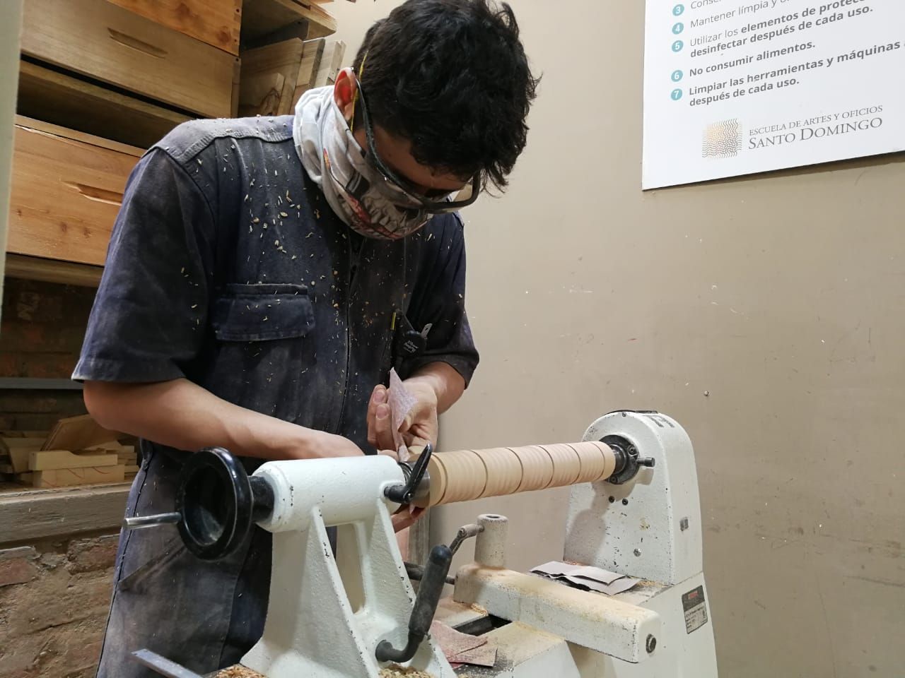 Persona en un taller torneando madera, con gafas de seguridad y mascarilla.