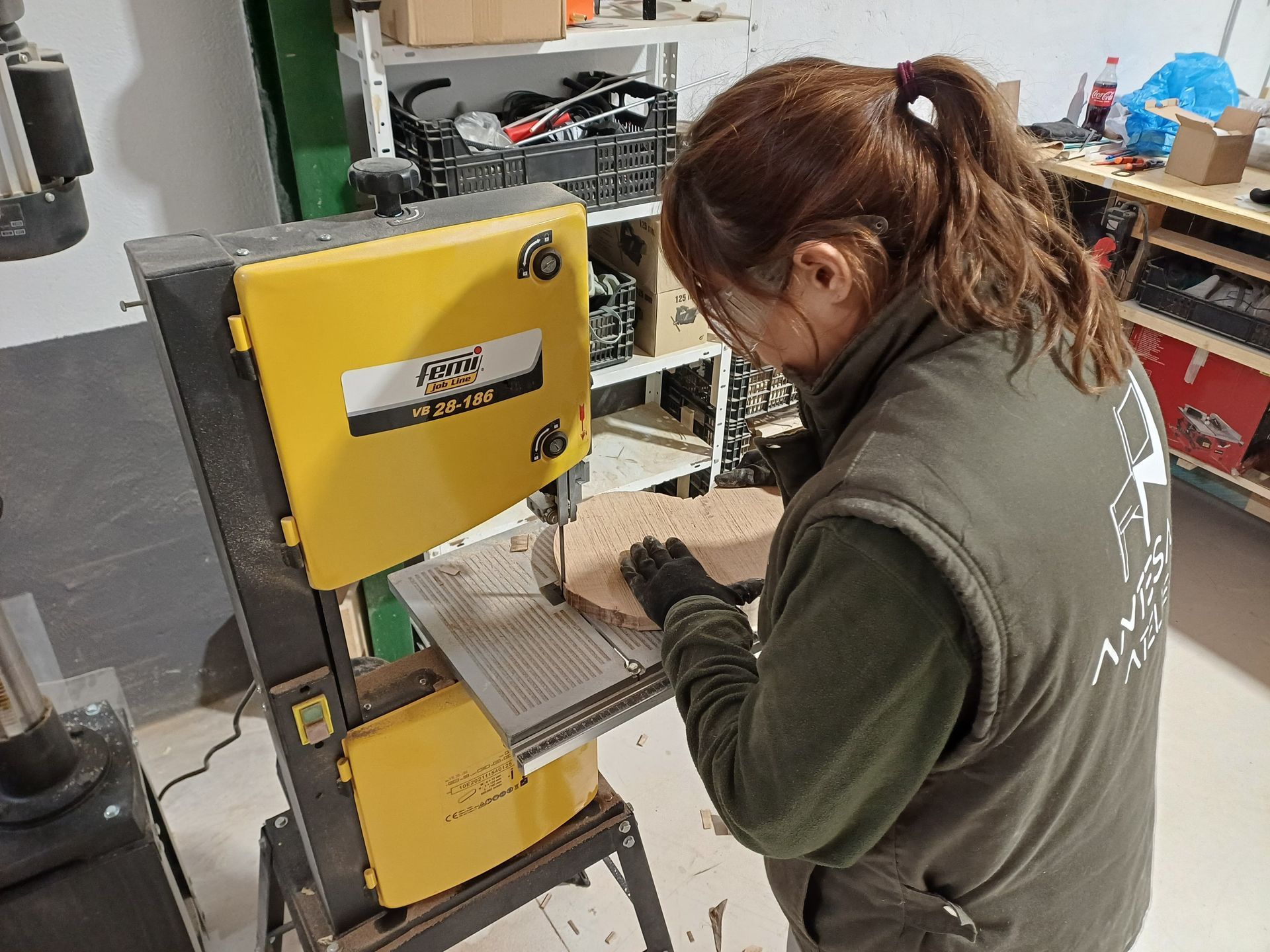 Mujer utilizando una sierra de cinta amarilla para cortar madera en un taller. Lleva chaleco y guantes.