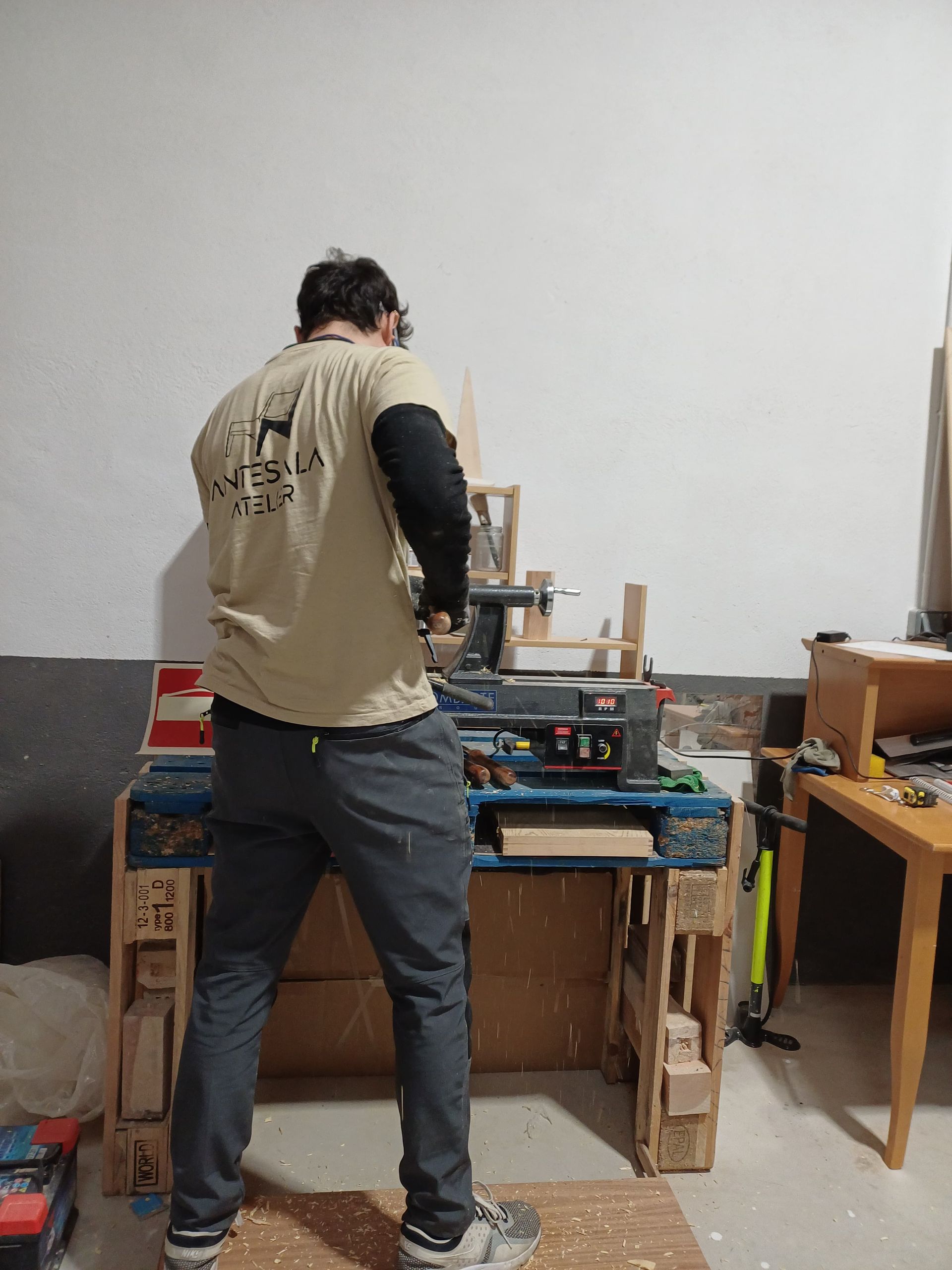 Hombre trabajando en un torno de madera, de pie en un taller. Camisa beige, pantalones grises. Banco de trabajo de madera