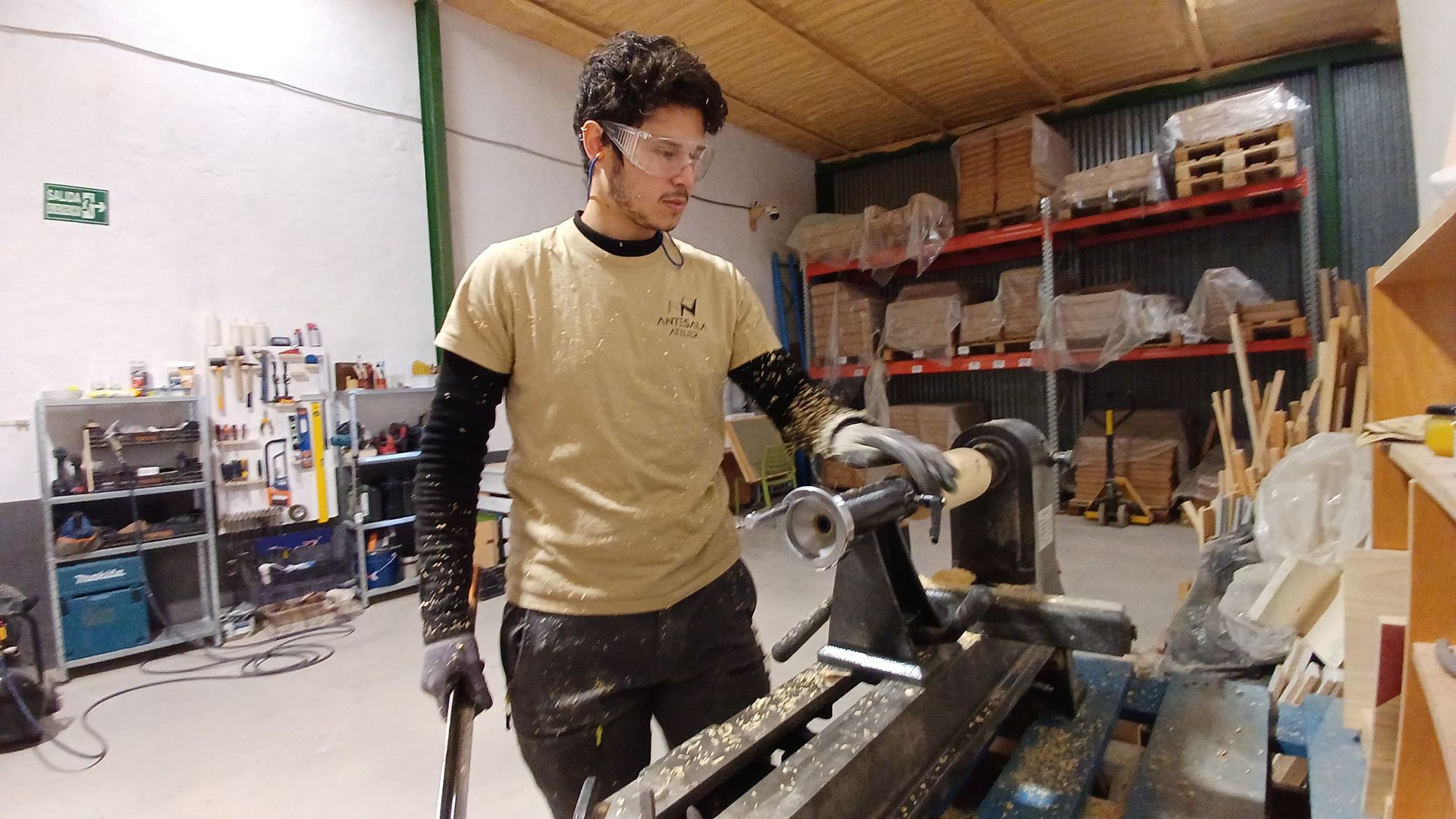 Hombre con gafas de seguridad y guantes trabajando en un torno de madera en un taller.