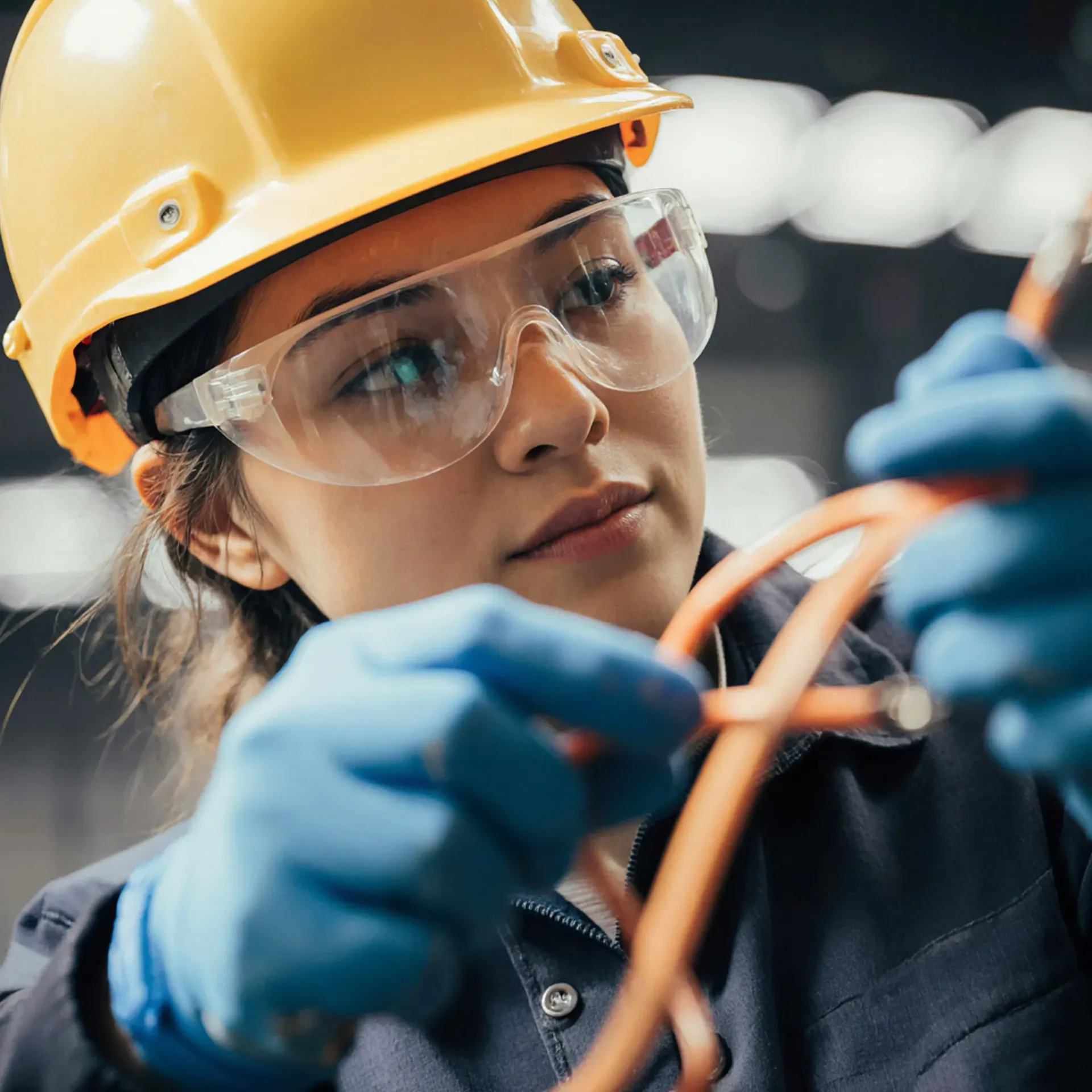 Una mujer con casco amarillo y gafas de seguridad examina un cableado naranja. Lleva guantes azules.