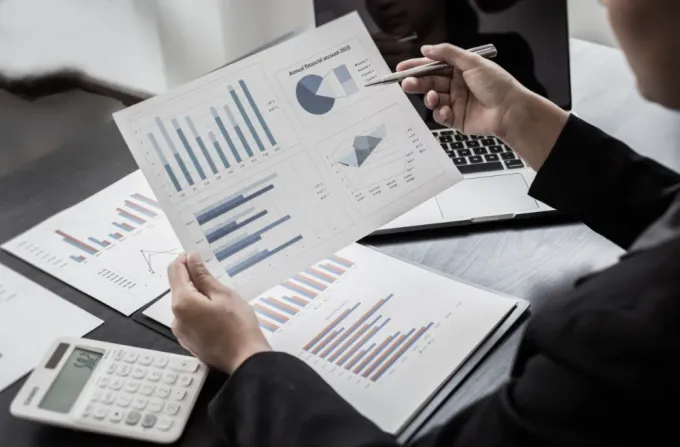 Persona de traje señala gráficos financieros, sentada en un escritorio con una computadora portátil y una calculadora.