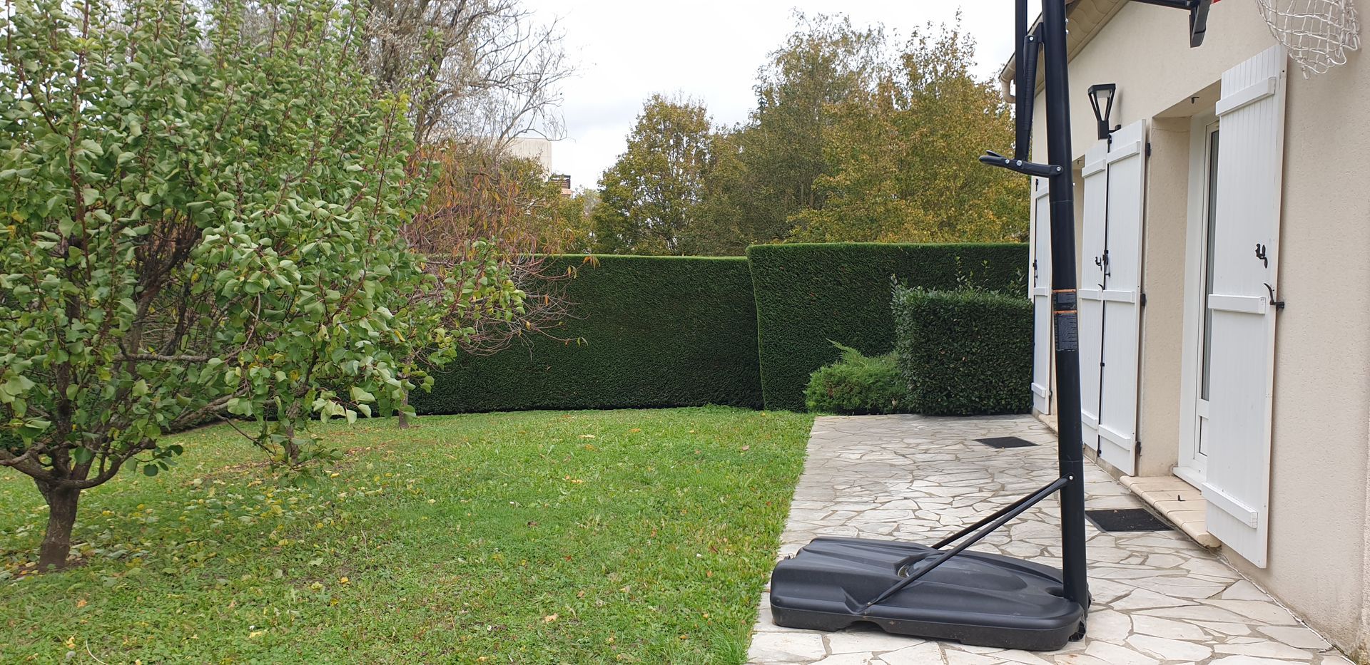Jardin avec pelouse verte, haies, arbres et terrasse de maison à côté d'un panier de basket.