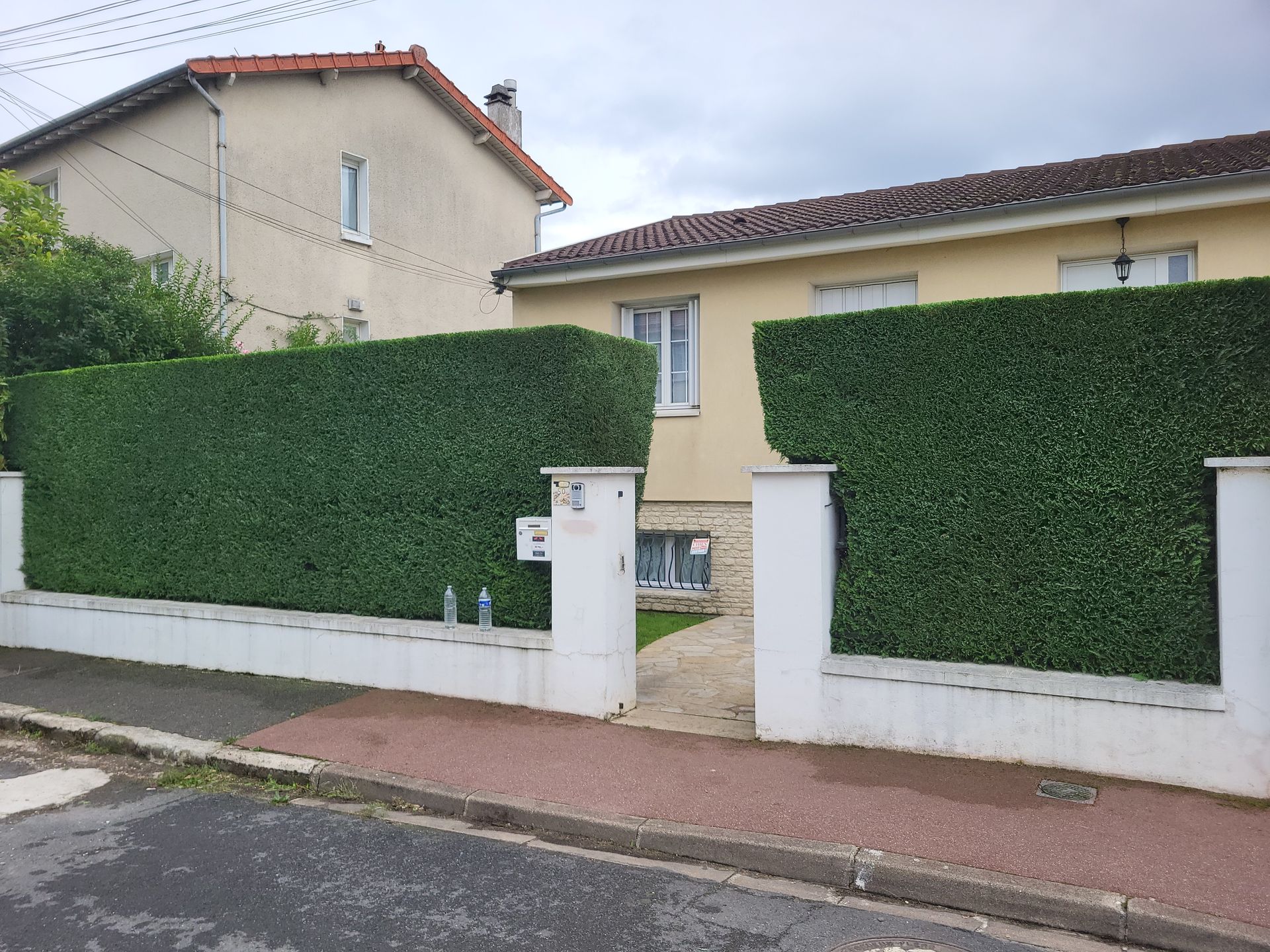 Maison d'habitation derrière de hautes haies vertes, avec un portail blanc et un trottoir devant.