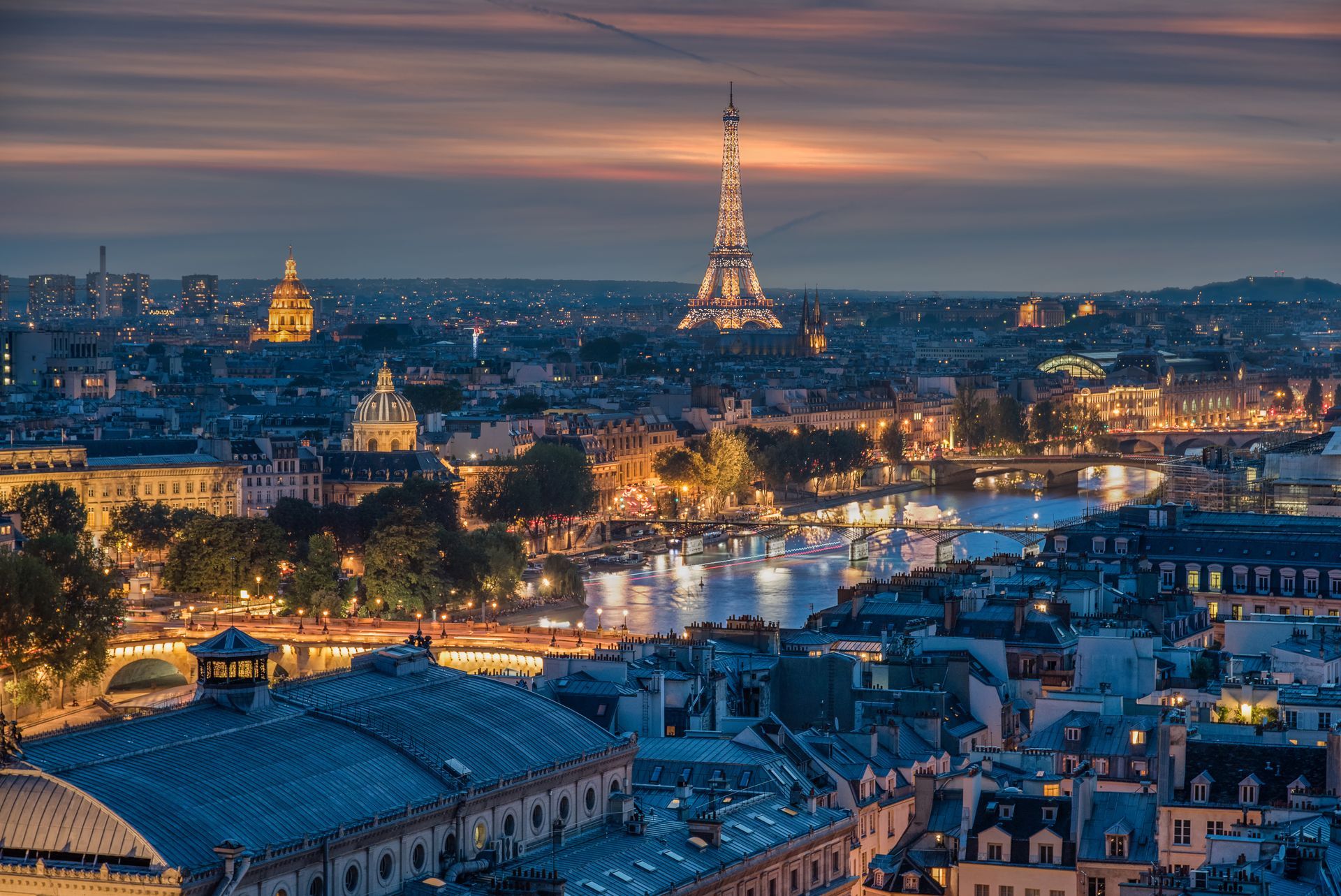 L'horizon parisien au crépuscule, avec la tour Eiffel qui brille au-dessus des lumières de la ville et des toits.