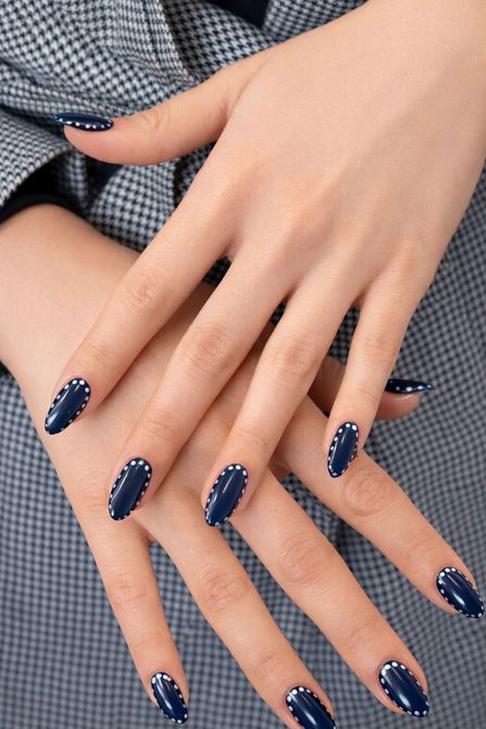 Manos con uñas almendradas de color azul oscuro, pequeñas joyas blancas en la base, apoyadas sobre una chaqueta gris.