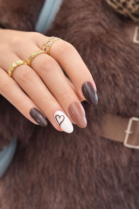 Mano con uñas almendradas en tonos marrones y nude, una de ellas con diseño de corazón, con aros dorados.