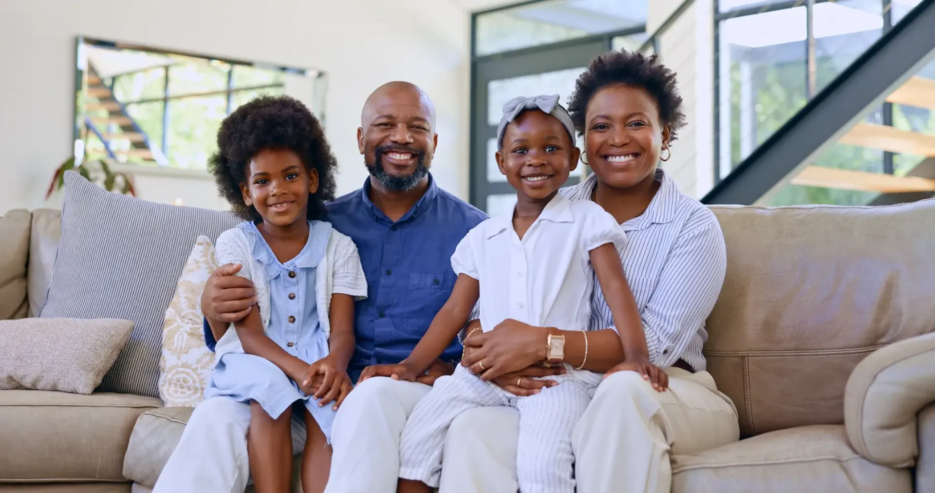 Family of four sitting together and smiling on a couch in a bright living room.