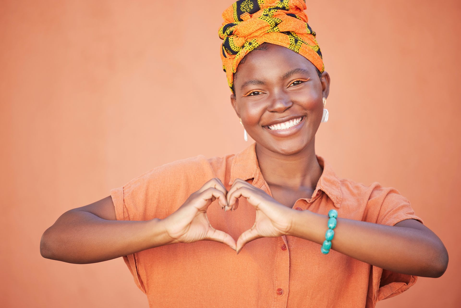Smiling person in orange shirt and headwrap making a heart shape with hands against a peach wall