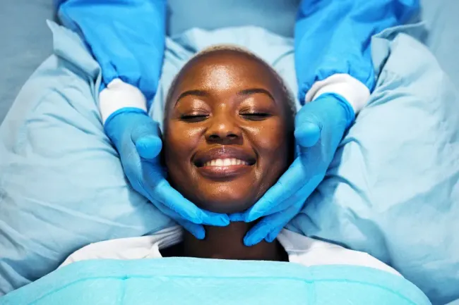 Person lying on a treatment bed with eyes closed while gloved hands cradle their face, smiling