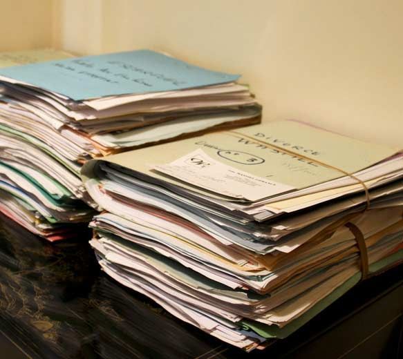 Des piles de papiers et de cahiers s'entassent sur un bureau dans une pièce encombrée.