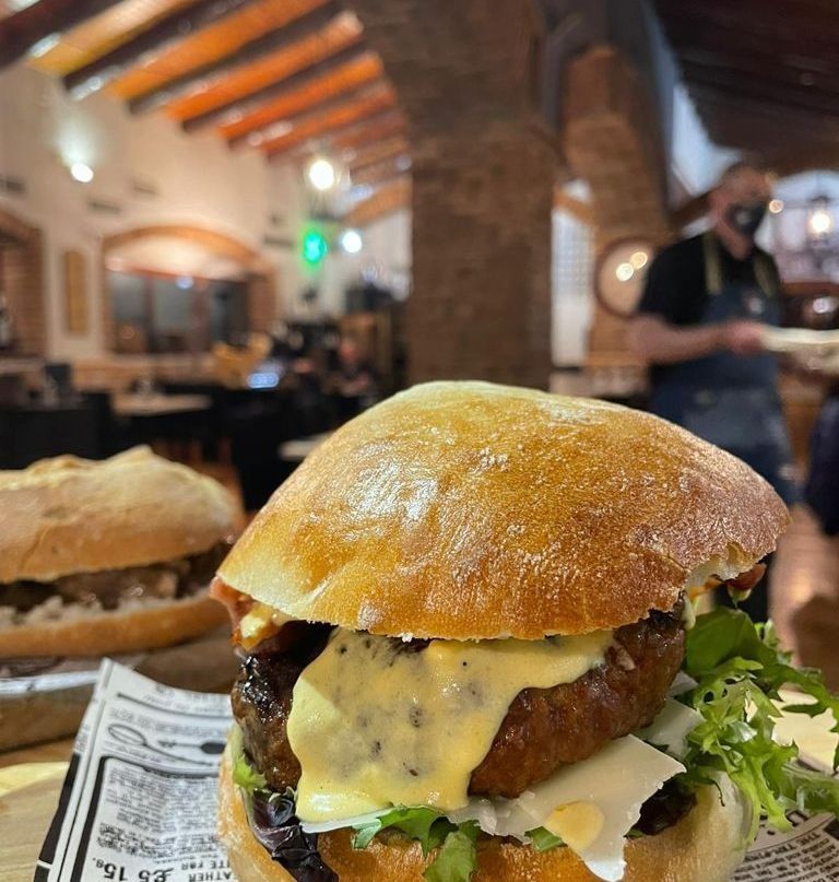 Hamburguesa sobre una superficie de madera en un restaurante. Se ve parcialmente otra hamburguesa al fondo, con una persona con mascarilla.