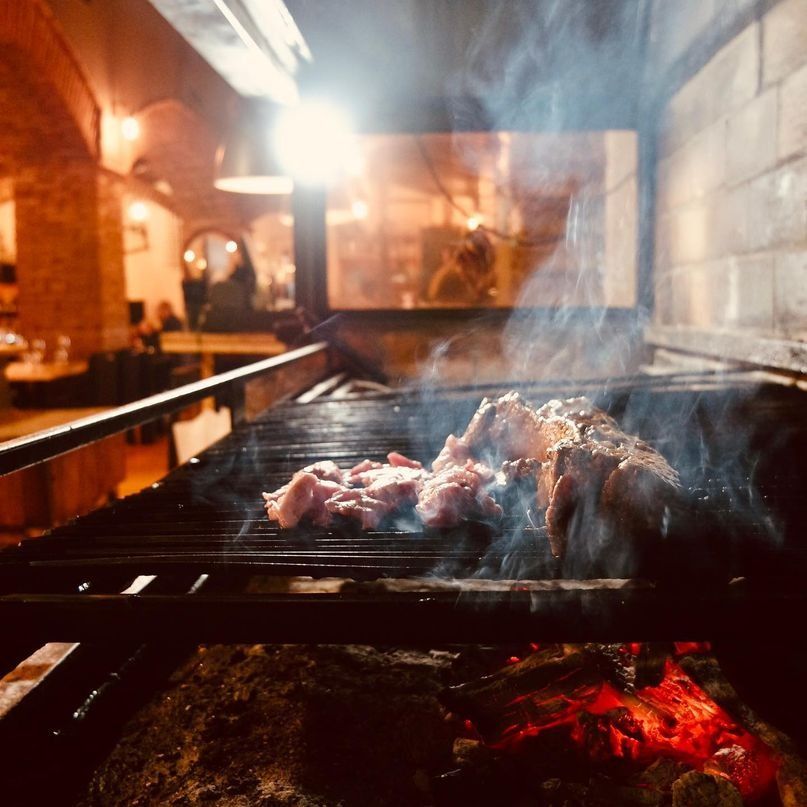 Carne asándose en una parrilla caliente, con humo saliendo. La escena se desarrolla en un restaurante con paredes de ladrillo y poca luz.