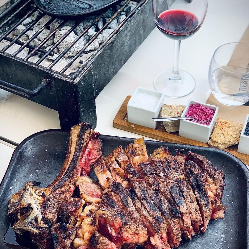 Bistec a la parrilla con una copa de vino tinto y salsas para mojar sobre una mesa blanca al lado de una pequeña parrilla.
