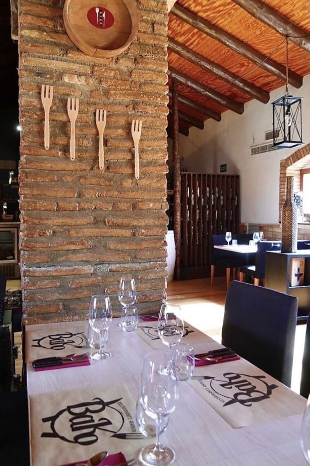 Interior del restaurante: mesa con vasos, cubiertos y manteles individuales. Pared de ladrillo con tenedores de madera decorativos. Al fondo, el comedor.