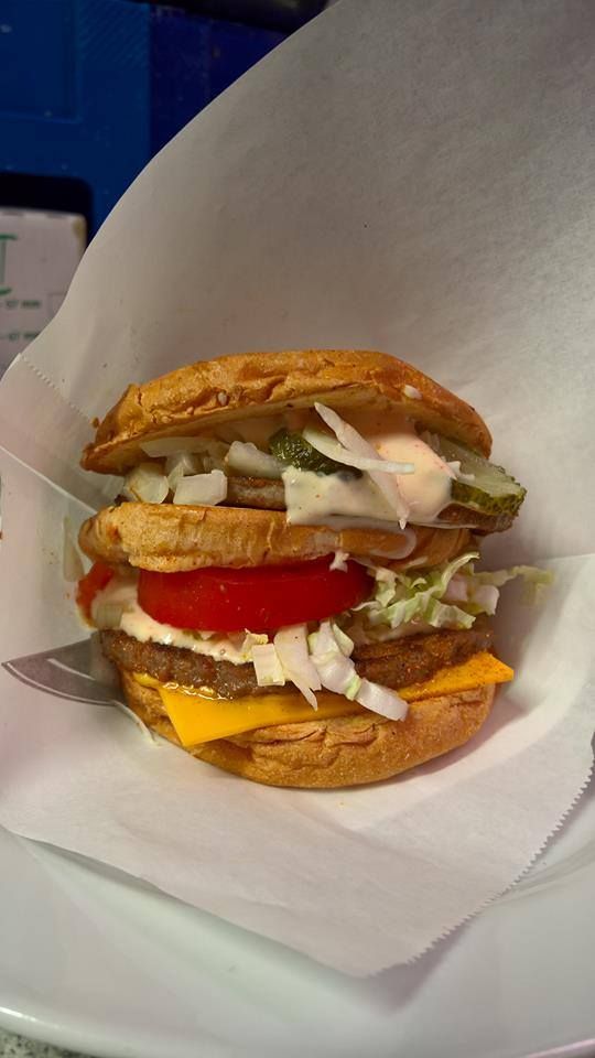 Kaksikerroksinen hampurilainen juustolla, tomaatilla, salaatilla, sipulilla, suolakurkuilla ja kastikkeella, kääritty valkoiseen paperiin.