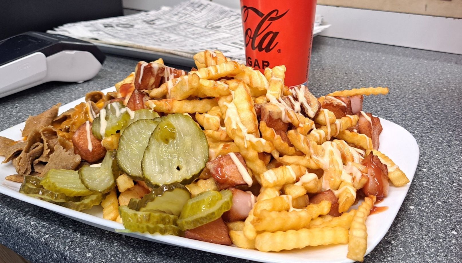 Lautanen, jossa on ranskalaisia, lihaa, suolakurkkuja ja kastiketta, punaisen Coca-Cola-mukin vieressä tiskillä.