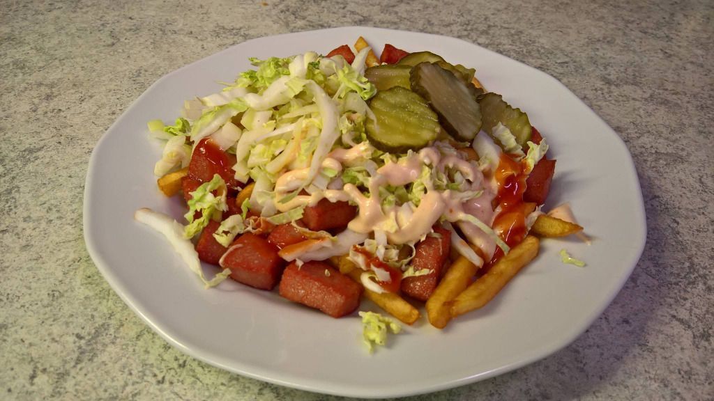 Ranskalaiset perunat, joiden päällä on viipaloitua makkaraa, suolakurkkuja, salaattia, ketsuppia ja kermakastiketta valkoisella lautasella.