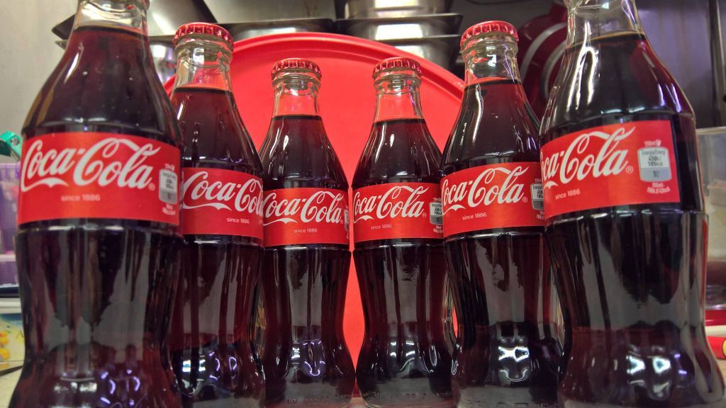 Kuusi lasista Coca-Cola-pulloa, joissa on punaiset etiketit ja korkit ja jotka on täytetty tummalla limsalla.
