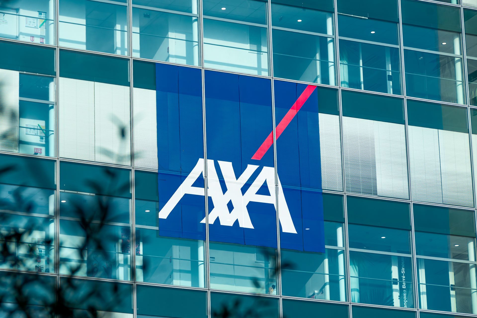 Le logo AXA sur un panneau bleu se reflète dans la façade vitrée d'un immeuble.