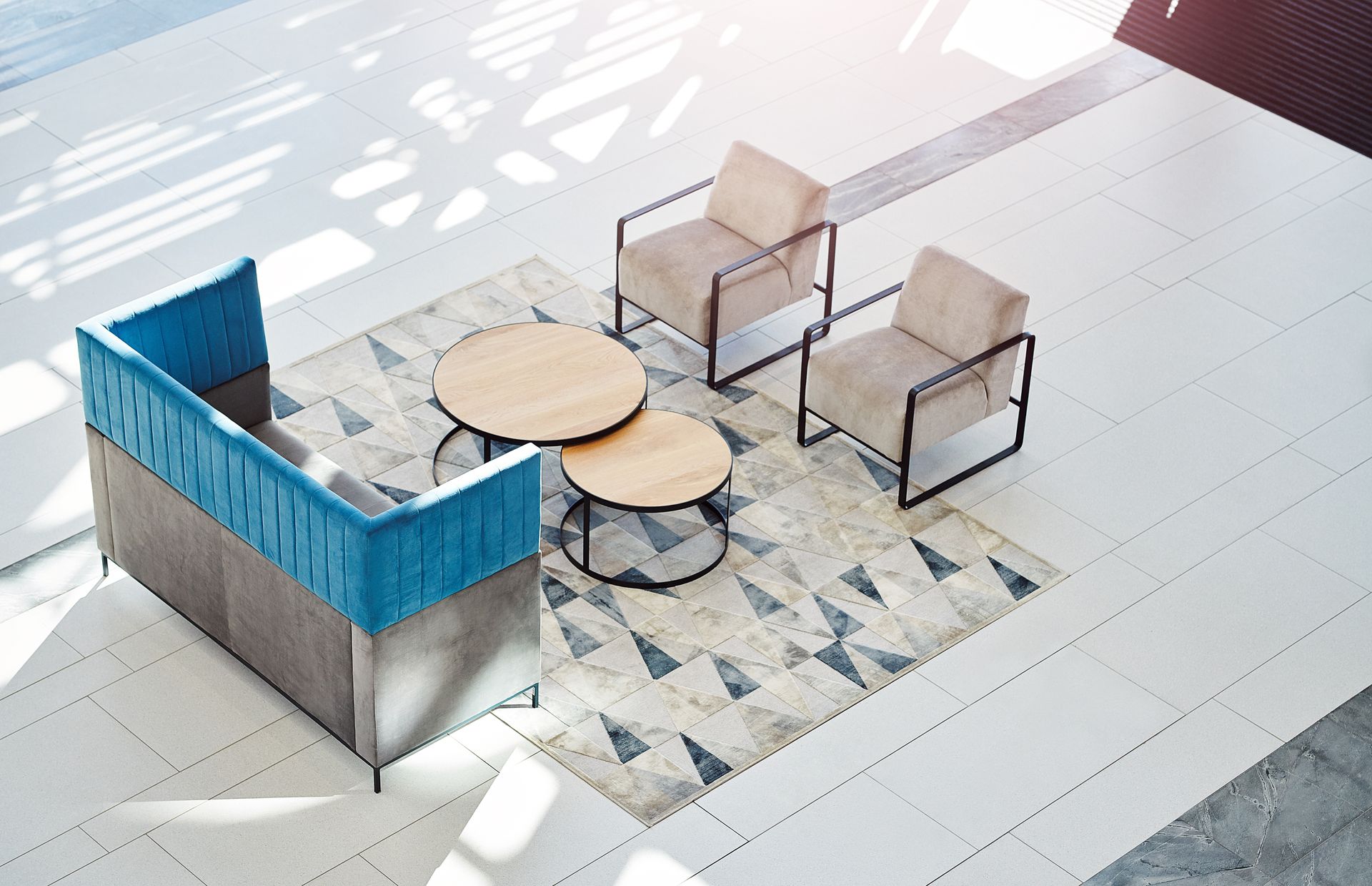 Vue aérienne d'un coin salon avec un canapé à dossier bleu, deux fauteuils beiges et deux tables rondes en bois sur un tapis à motifs.