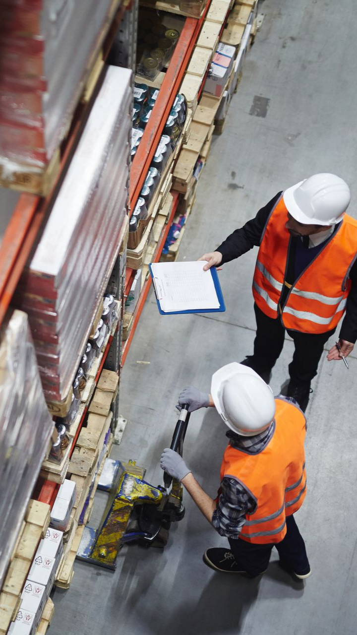 Deux magasiniers, vêtus de gilets haute visibilité et de casques de chantier, examinent des documents près de rayonnages à palettes.