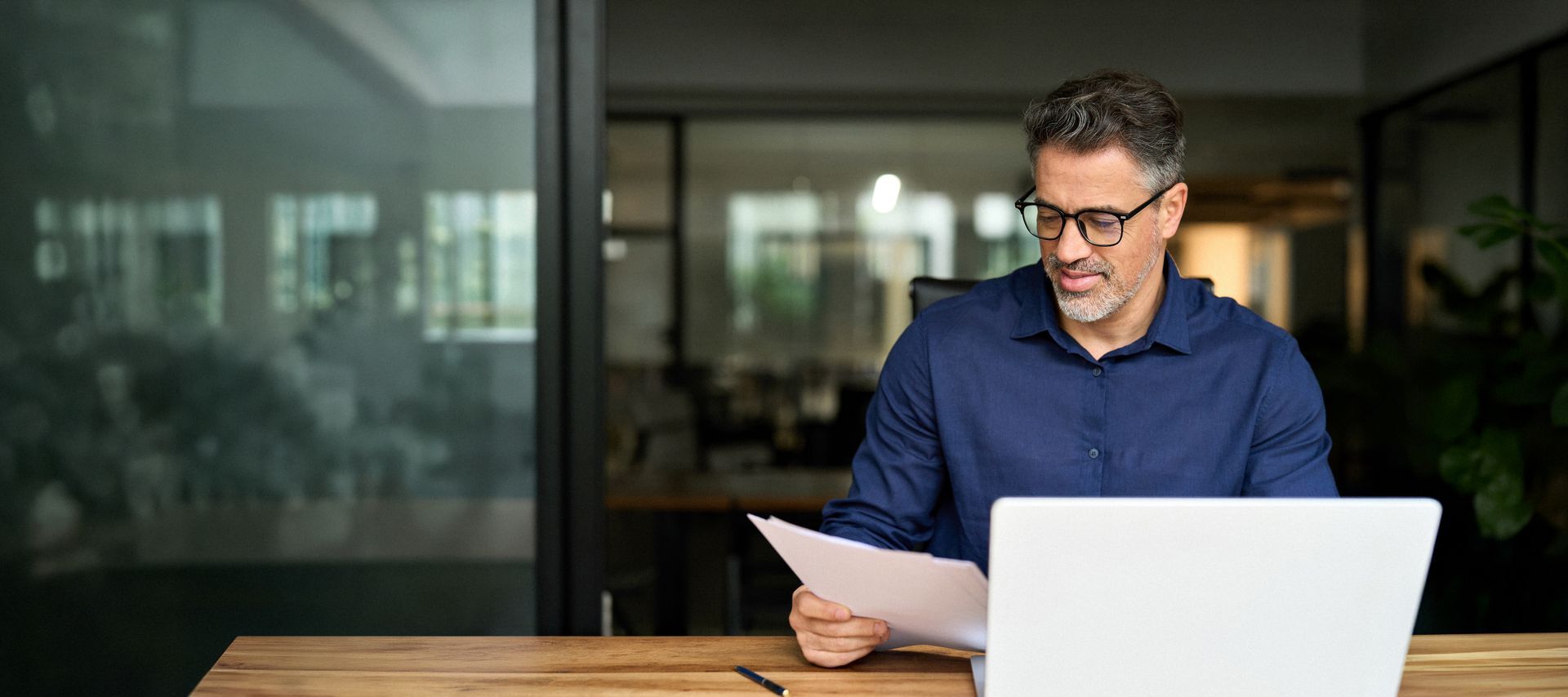 Un homme portant des lunettes lit des documents sur un ordinateur portable dans un bureau moderne.