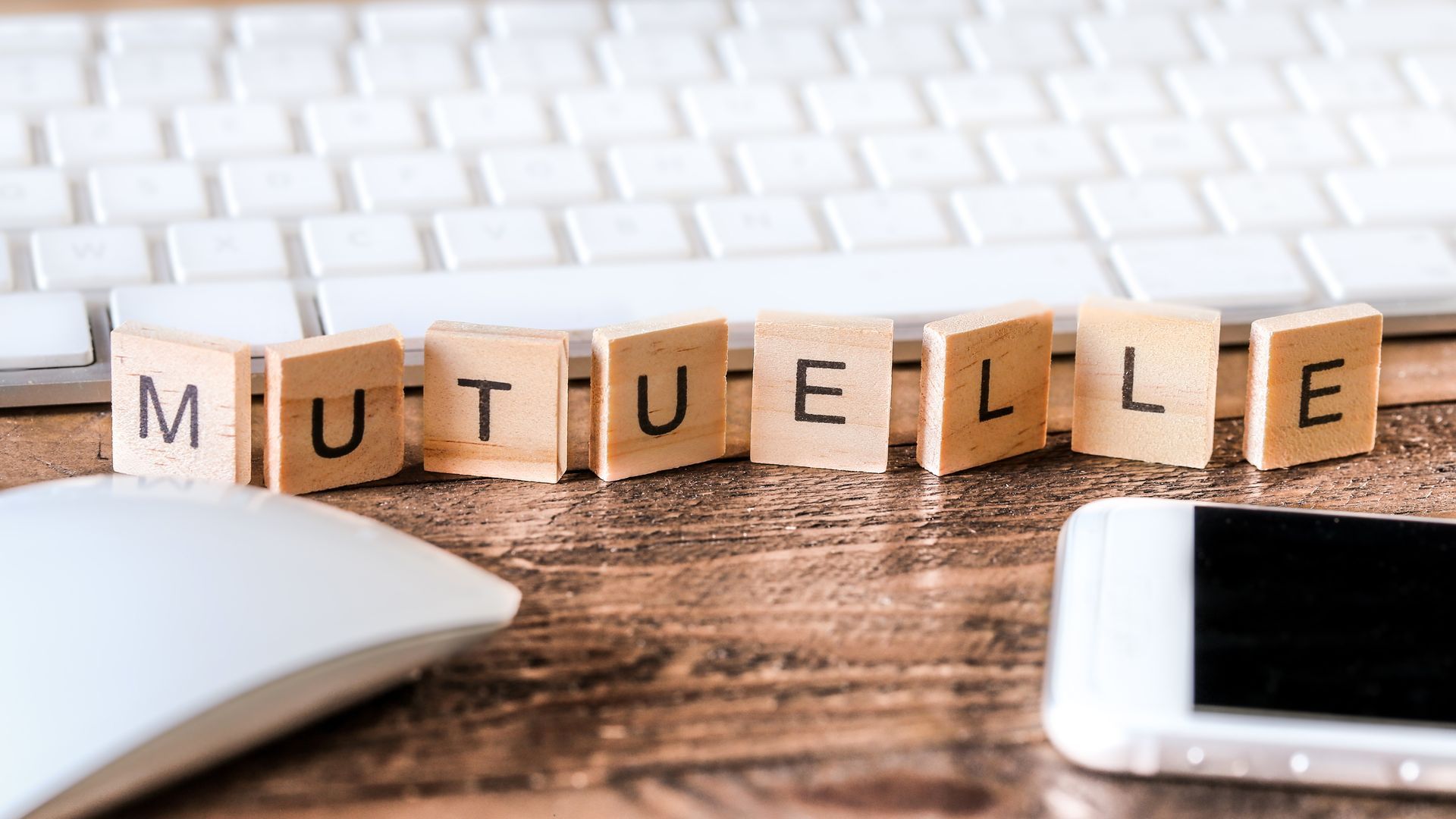 Des lettres en bois formant le mot MUTUELLE sont posées sur un bureau, à côté d'un clavier d'ordinateur et d'un smartphone.