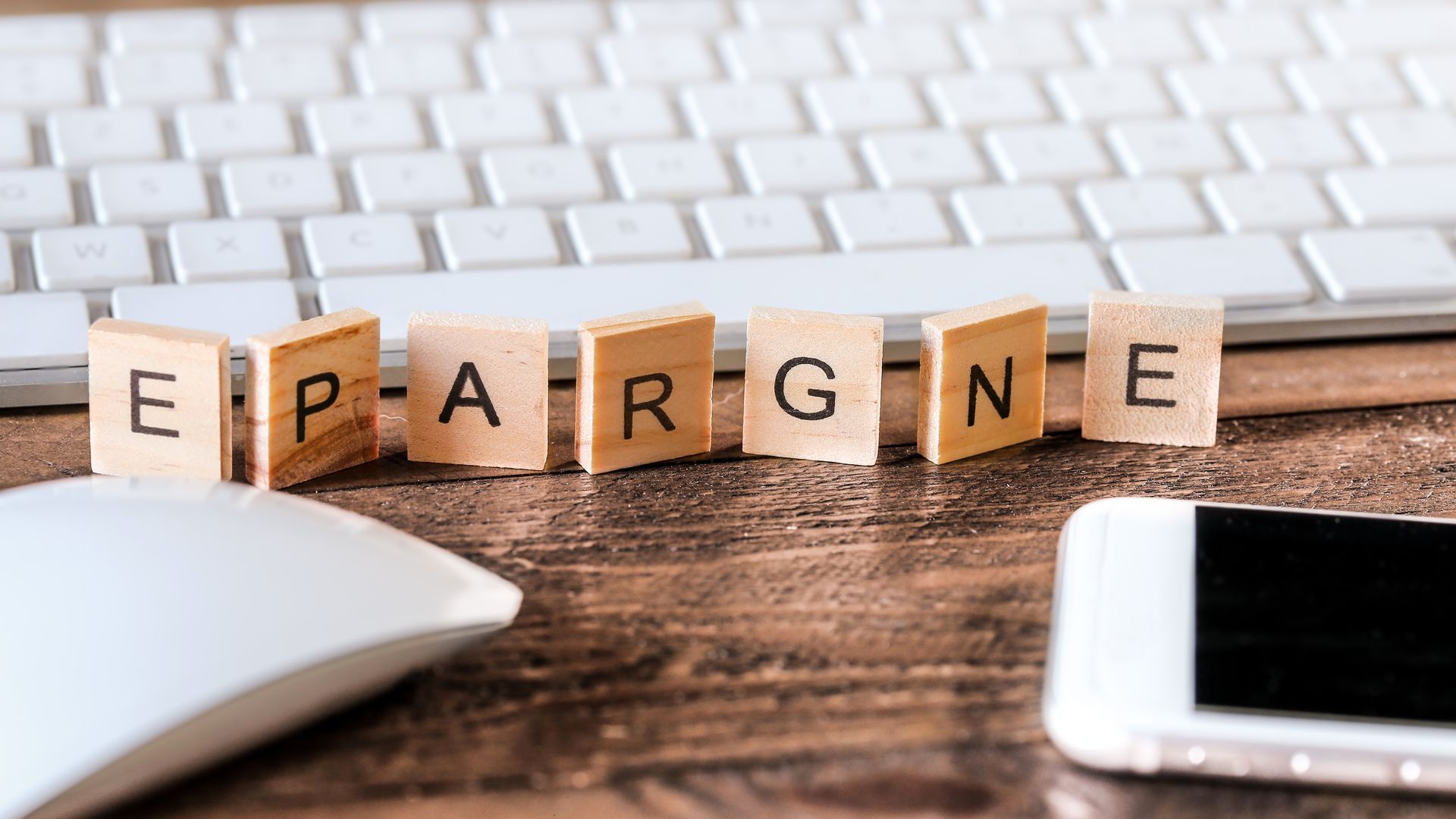 Des lettres en bois formant le mot  EPARGNE  sont disposées sur un bureau en bois, avec un clavier blanc et un smartphone.