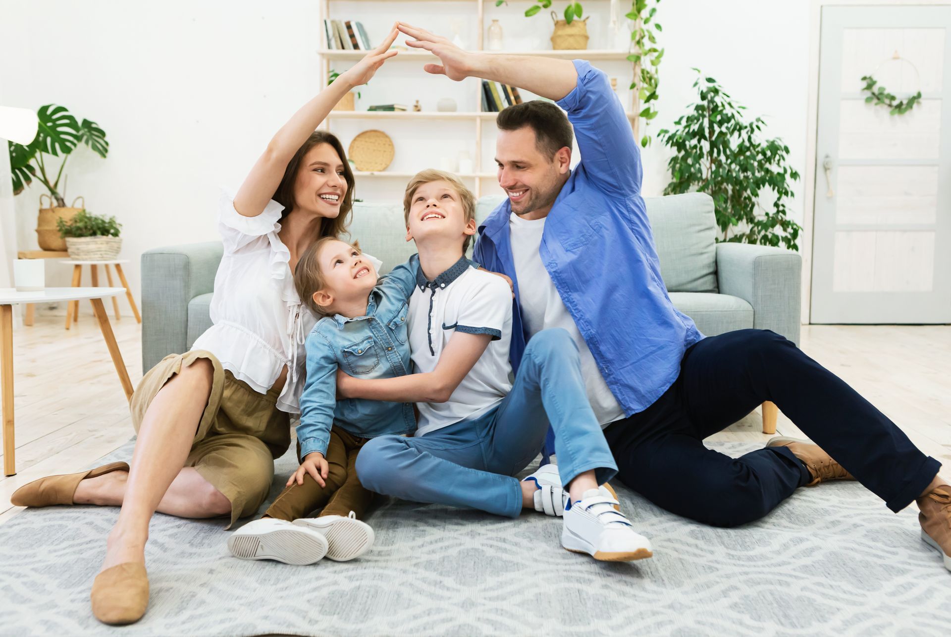 Une famille souriante est assise sur un tapis de salon, formant un toit au-dessus de leurs têtes avec leurs bras.