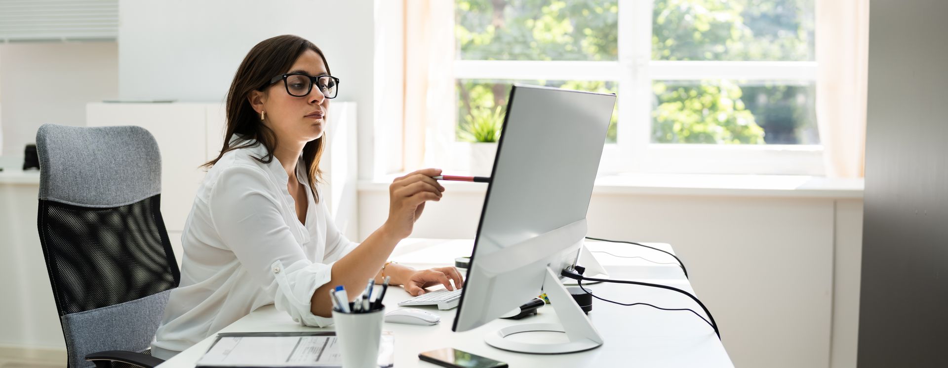 Dans un bureau lumineux, une personne vêtue d'une chemise blanche et portant des lunettes est assise à un bureau, pointant du doigt un écran d'ordinateur avec un stylo.