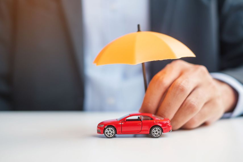 Une main gantée d'un costume tient un parapluie jaune miniature au-dessus d'une petite voiture jouet rouge posée sur une surface blanche.