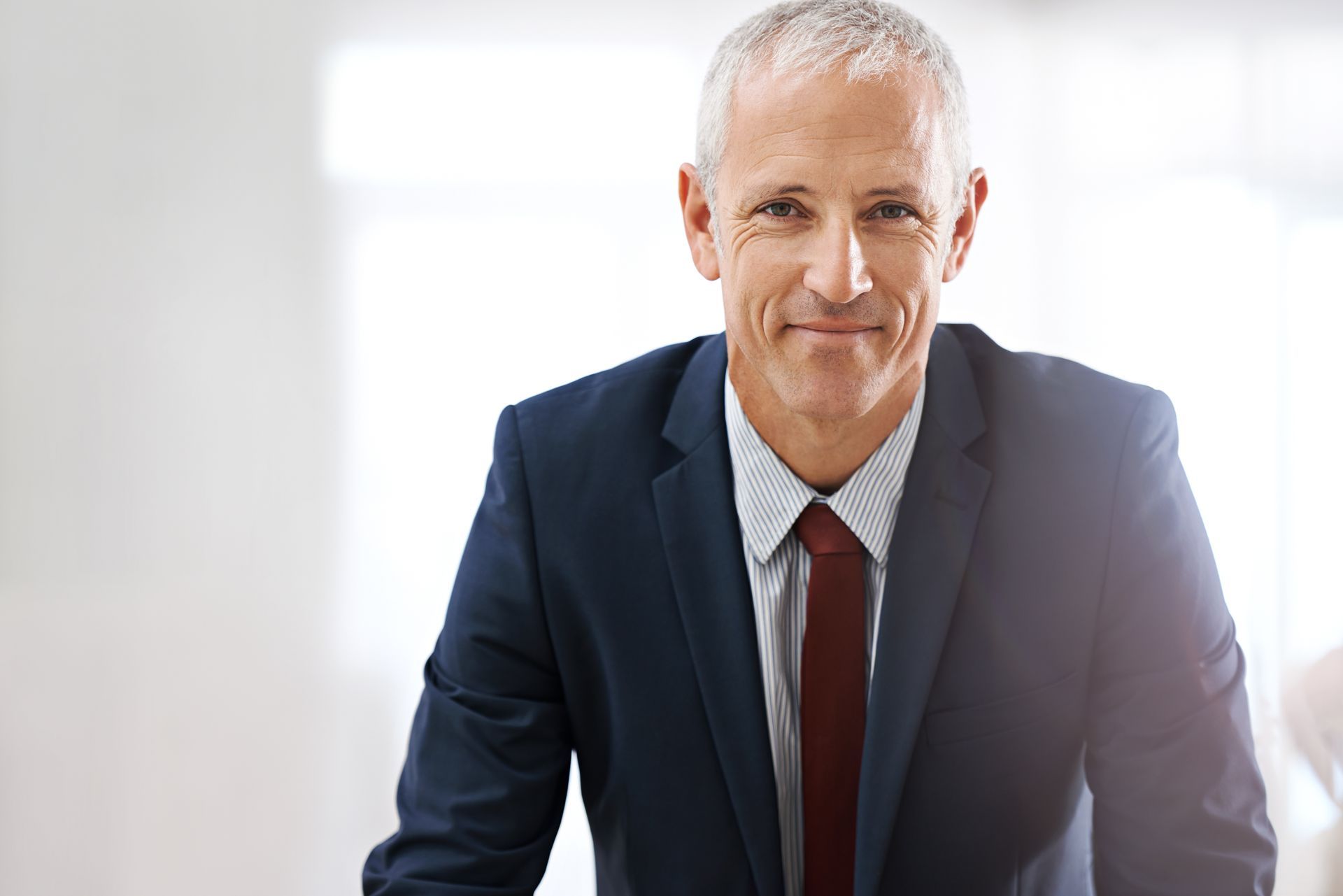 Un professionnel en costume bleu marine, chemise à rayures et cravate bordeaux sourit à l'objectif dans un bureau moderne et lumineux.
