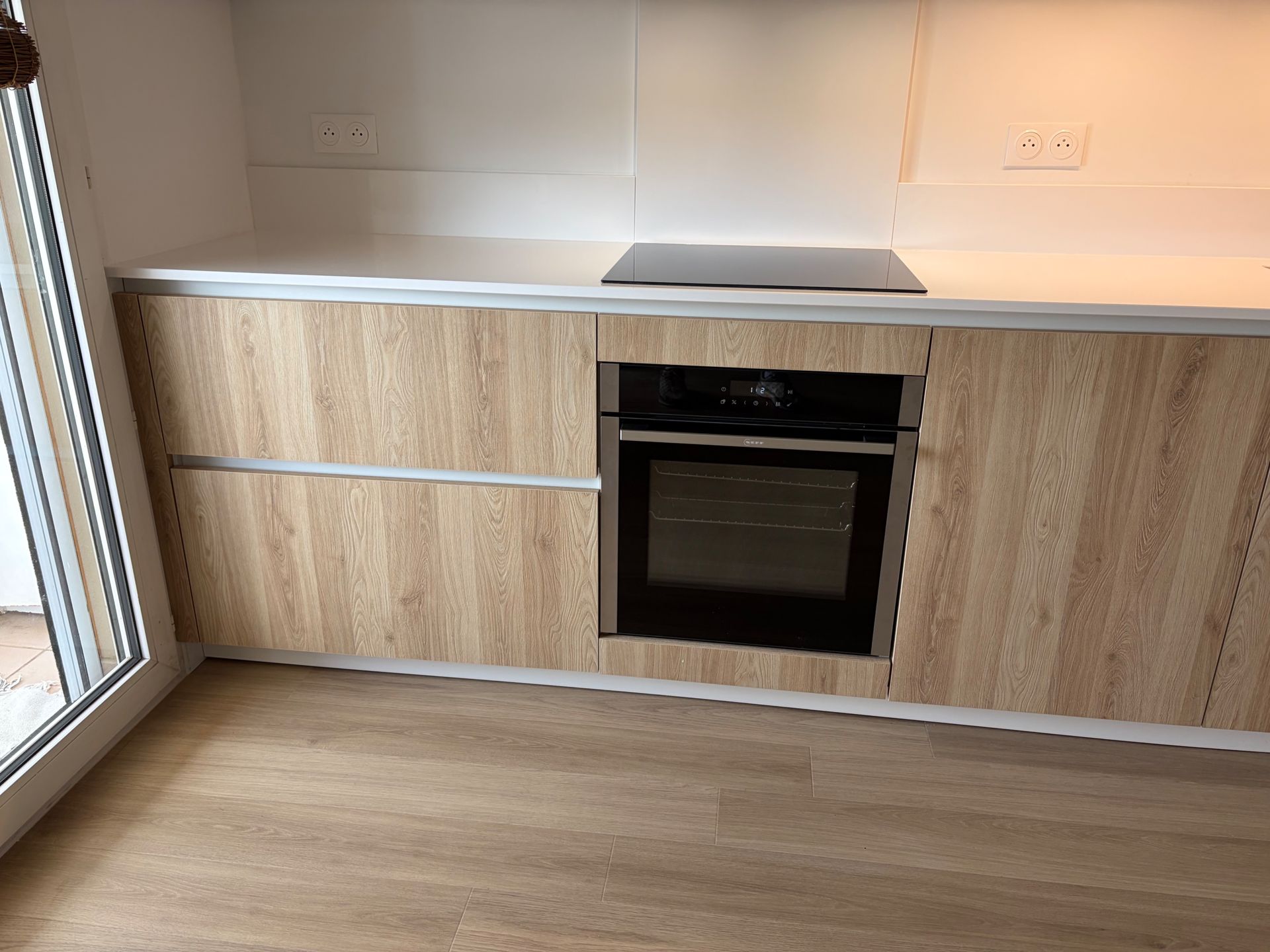 Cuisine moderne avec des placards en bois, un plan de travail blanc, un four encastré et un parquet en bois près d’une fenêtre