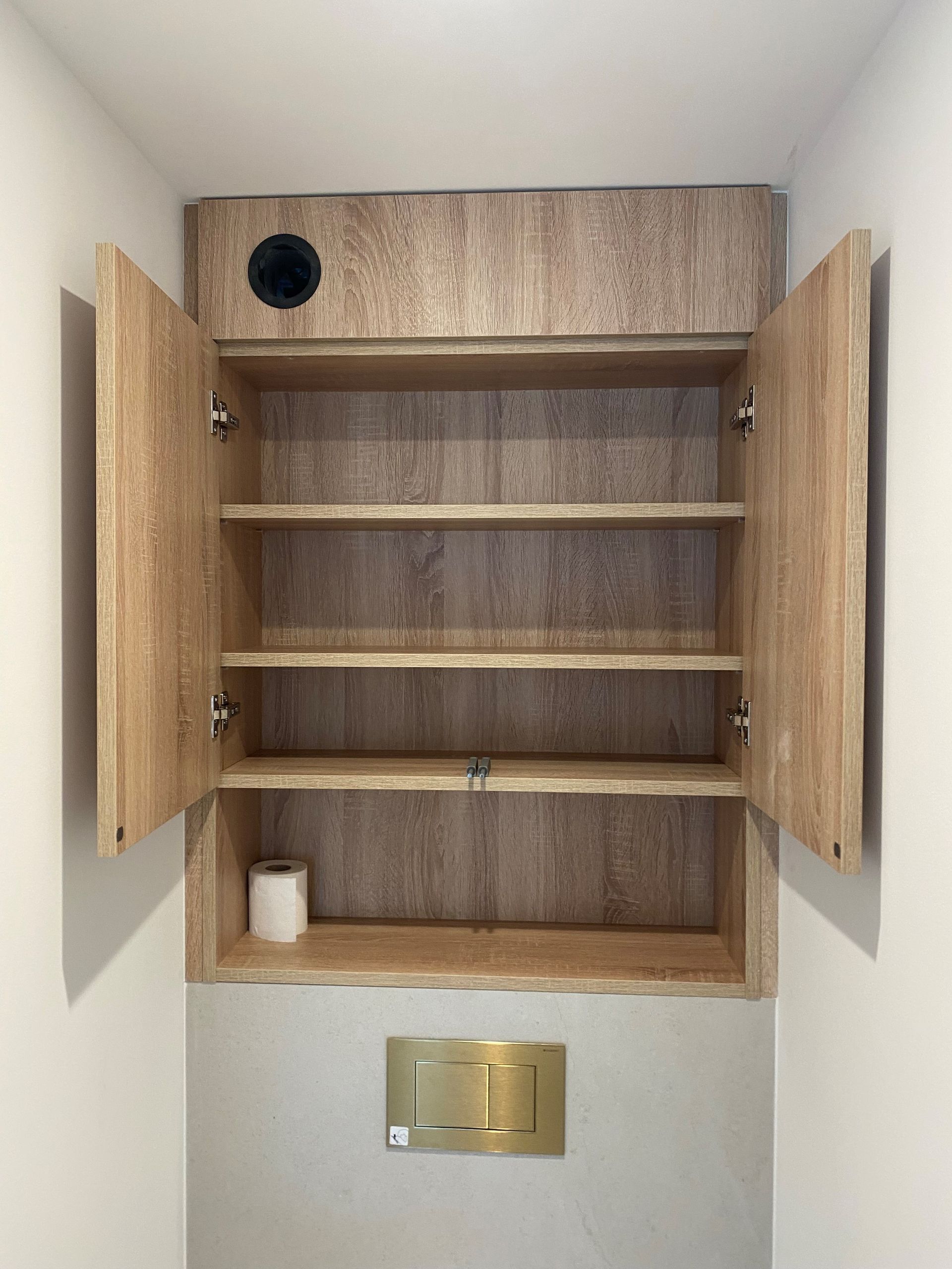 Meuble ouvert en bois avec étagères dans une salle de bains, au-dessus des toilettes. Un rouleau de papier toilette y est posé.