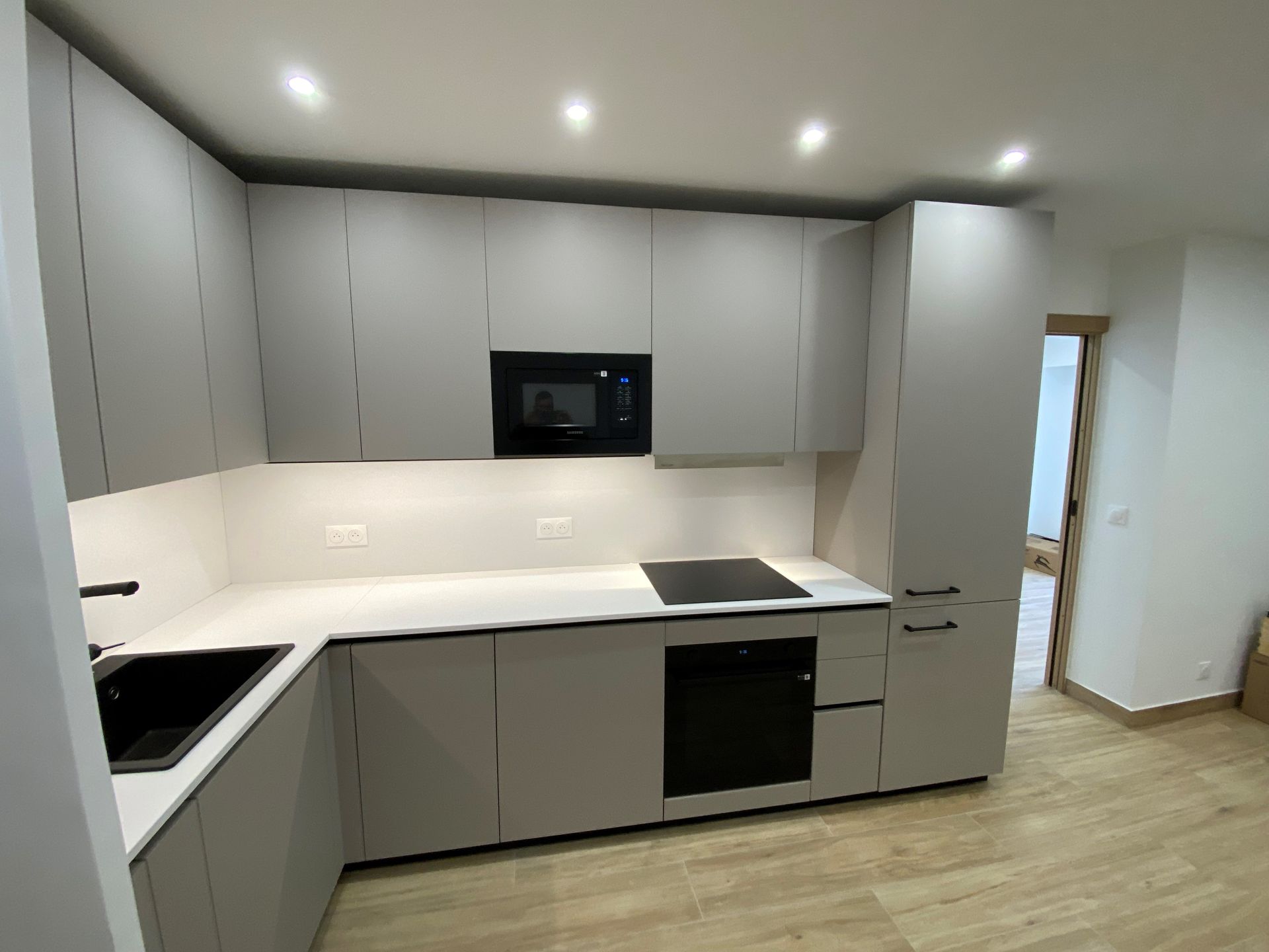 Cuisine grise moderne avec appareils électroménagers noirs, comptoirs blancs et parquet.