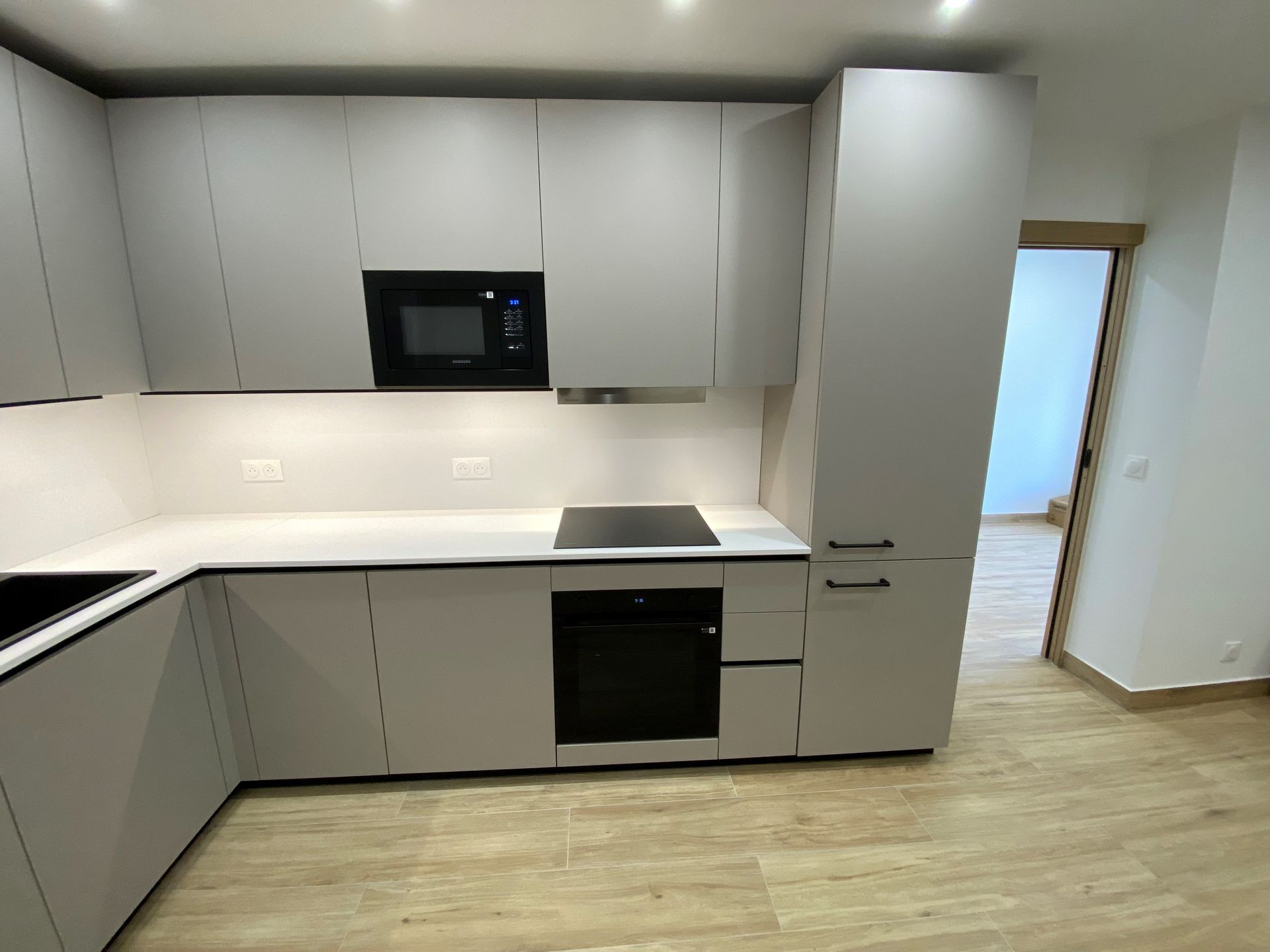 Cuisine moderne gris clair avec comptoirs blancs, appareils électroménagers noirs et une porte menant à une autre pièce.