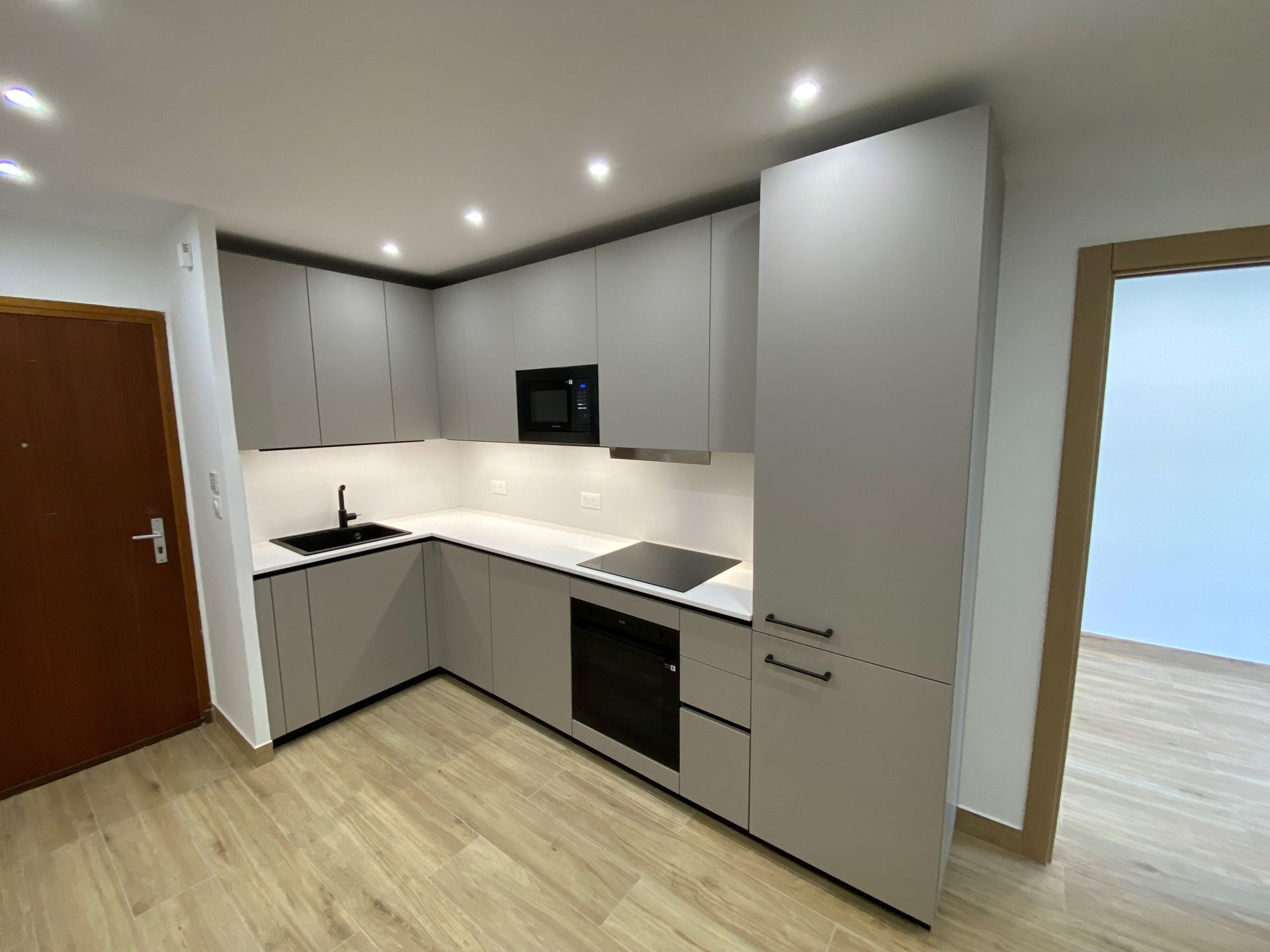 Cuisine grise moderne avec comptoirs blancs, appareils électroménagers noirs et parquet en bois clair.