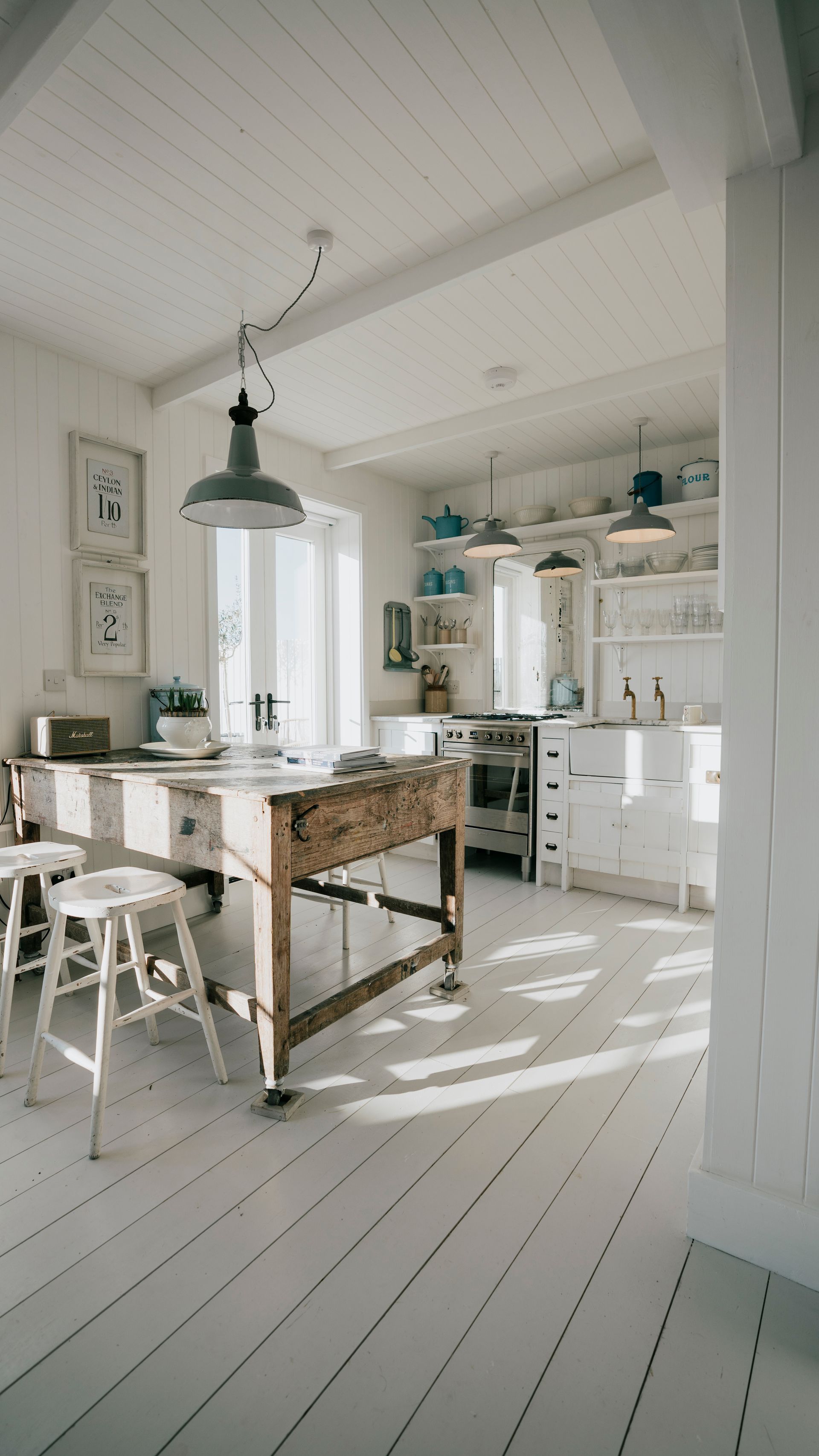 Cuisine de ferme d’un blanc éclatant, avec une table et des chaises en bois, une suspension et la lumière du soleil sur le sol peint